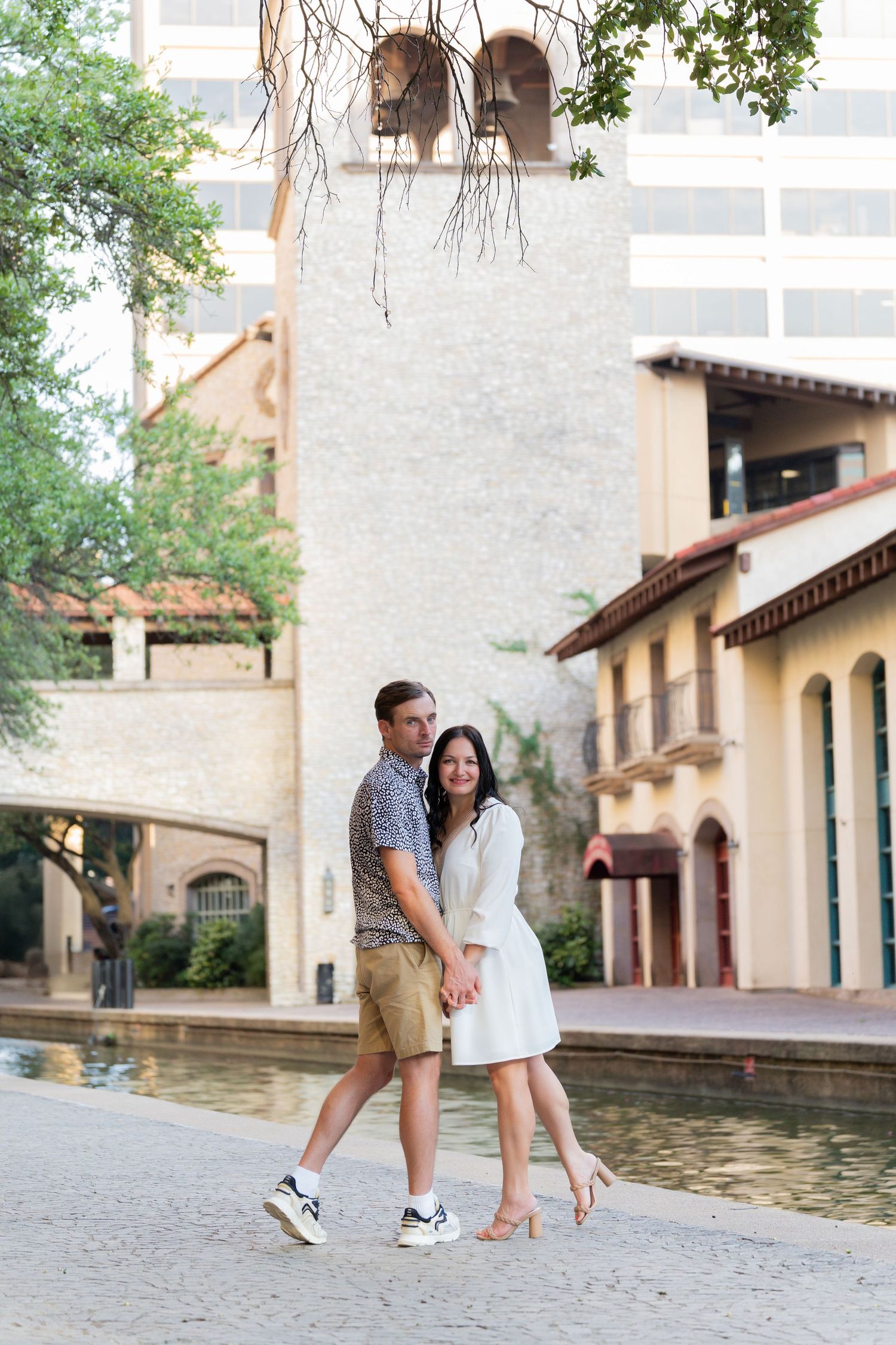 Playful couple interacts near Mediterranean-style architecture and a reflecting pool.