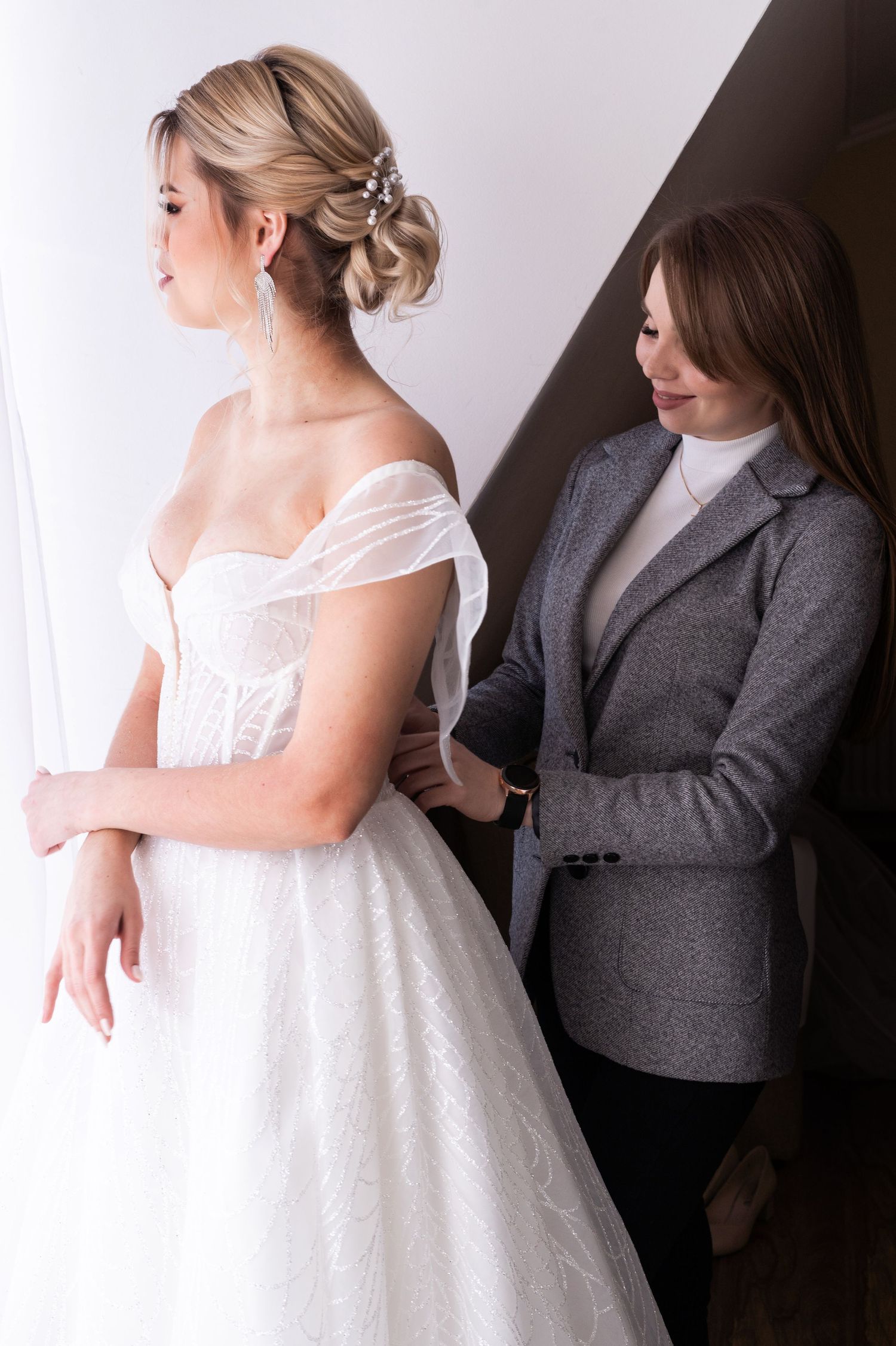 A bride in a white wedding dress gets help with final adjustments from someone wearing a grey suit.