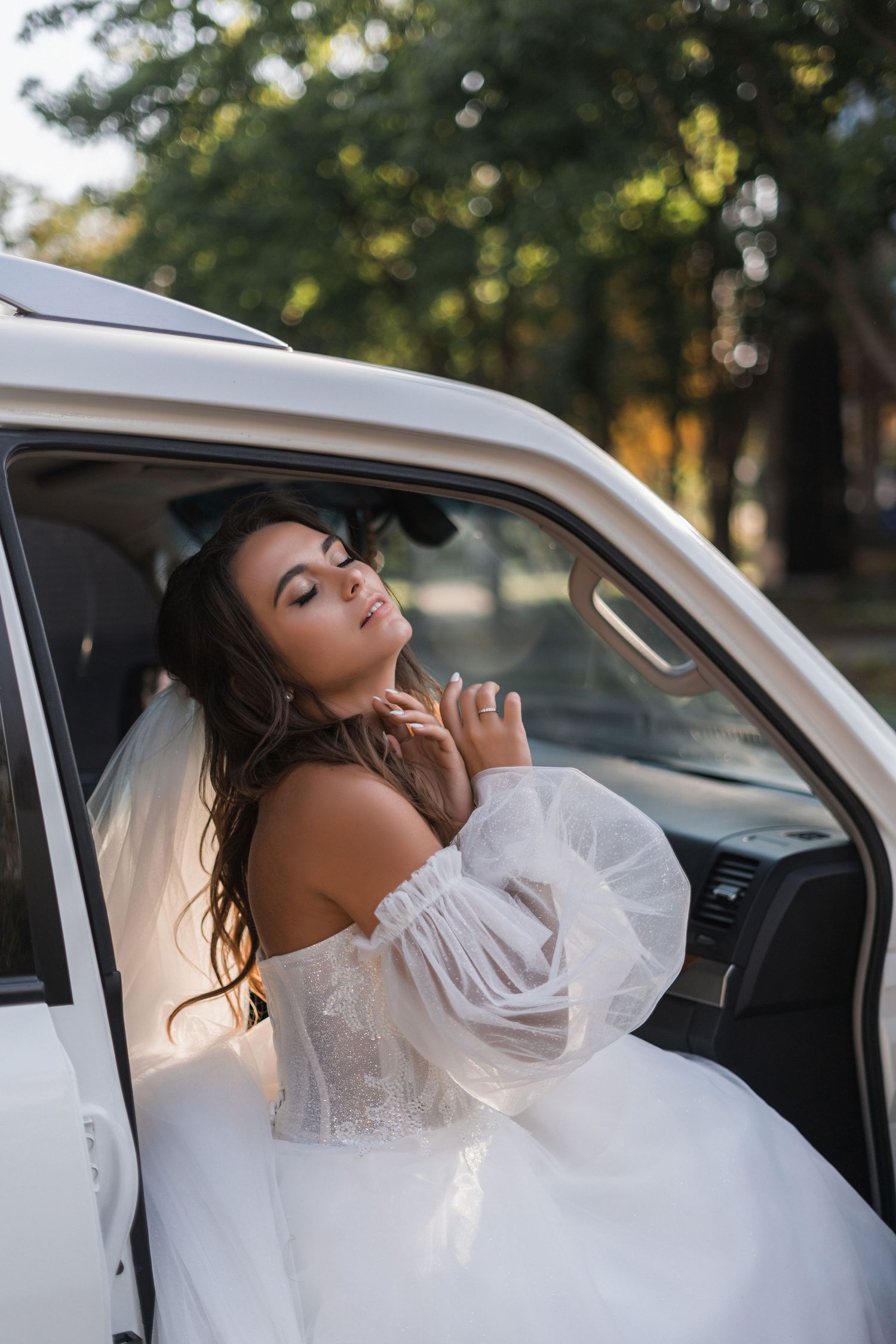 A bride in a white off-shoulder wedding dress poses elegantly in a white car during a golden hour photoshoot.