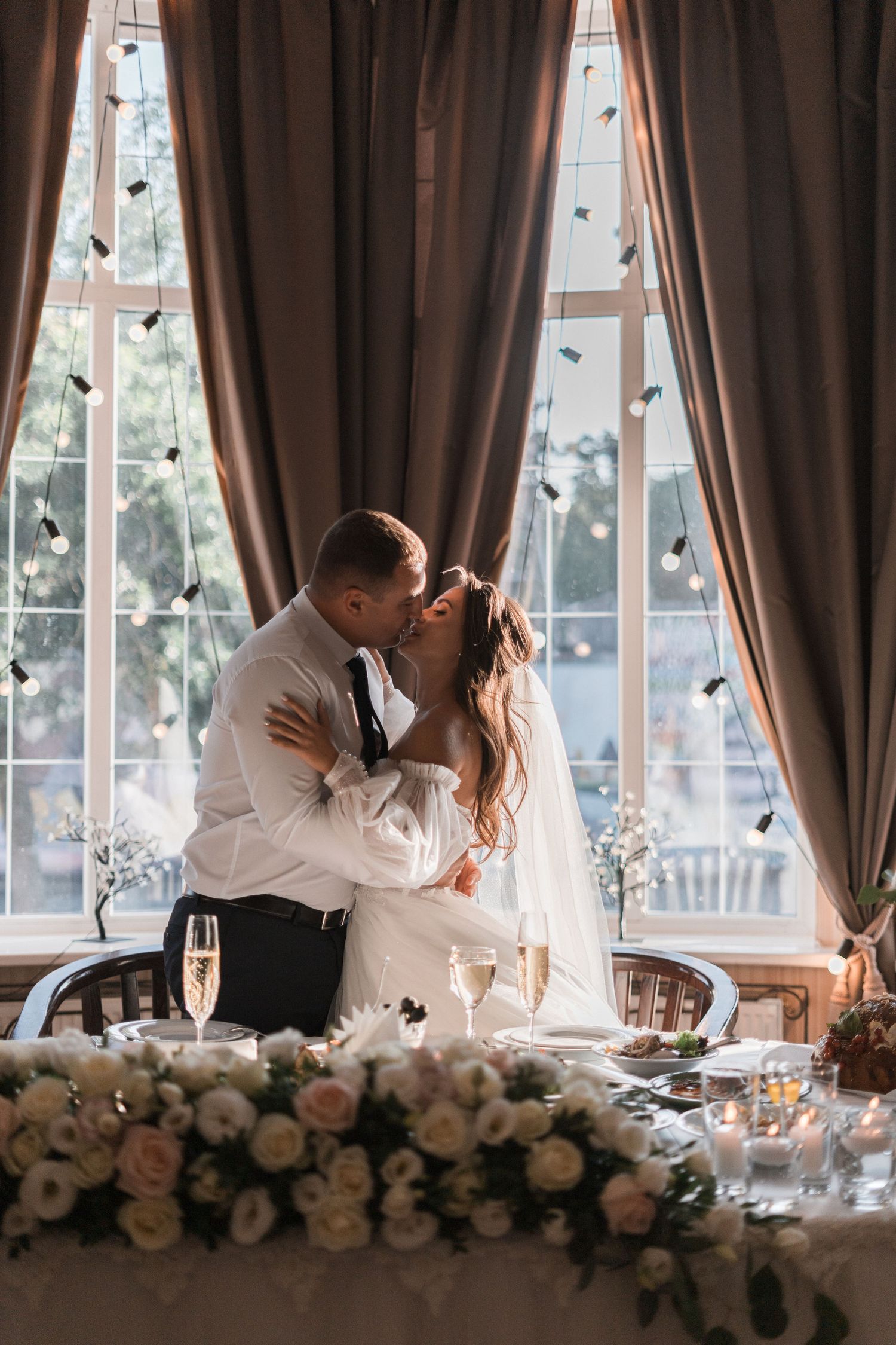Romantic wedding couple share an intimate kiss by elegant windows with floating butterflies and floral table decorations.