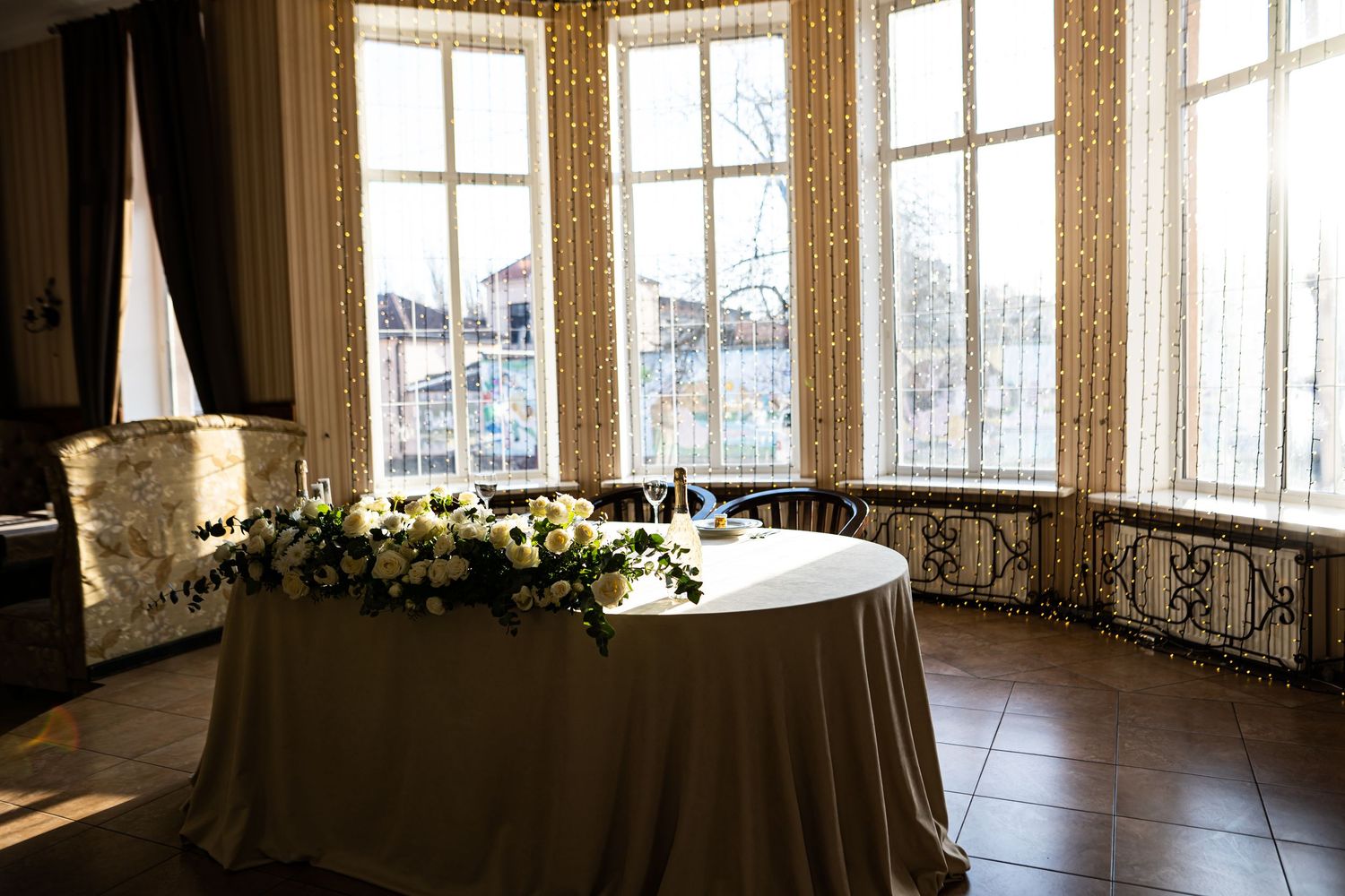 Elegant round table with floral centerpiece in sunlit corner room with ornate windows and city views.