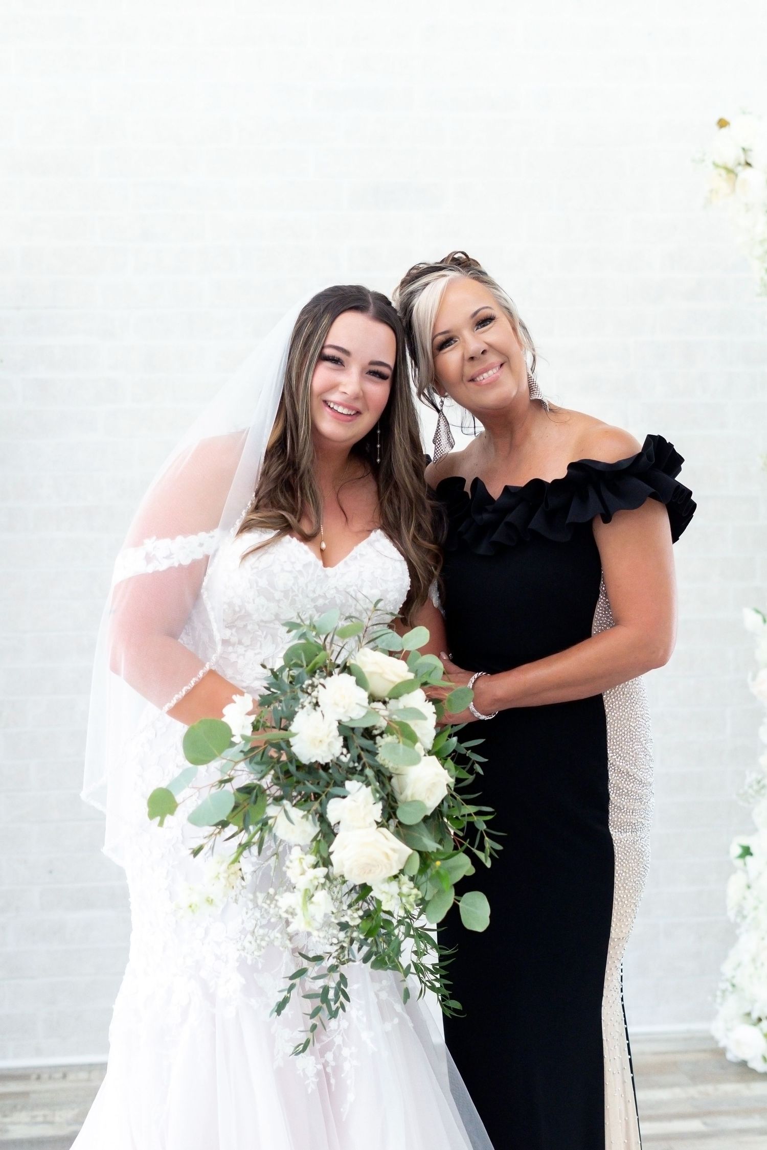 A bride in white dress with floral bouquet poses with companion in elegant black gown against white wall.