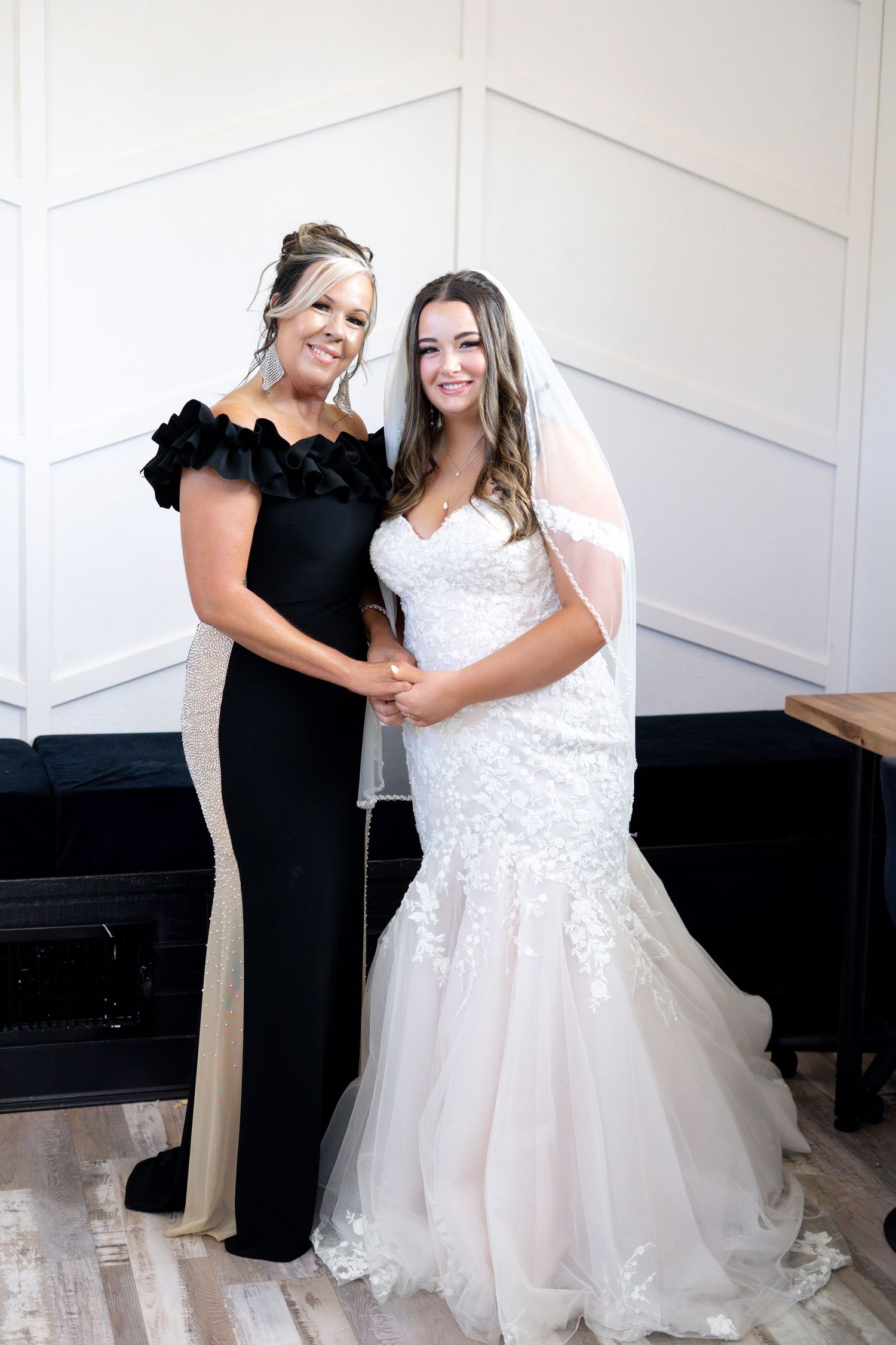 Two women embrace and smile while one wears a white wedding dress and the other wears black attire.