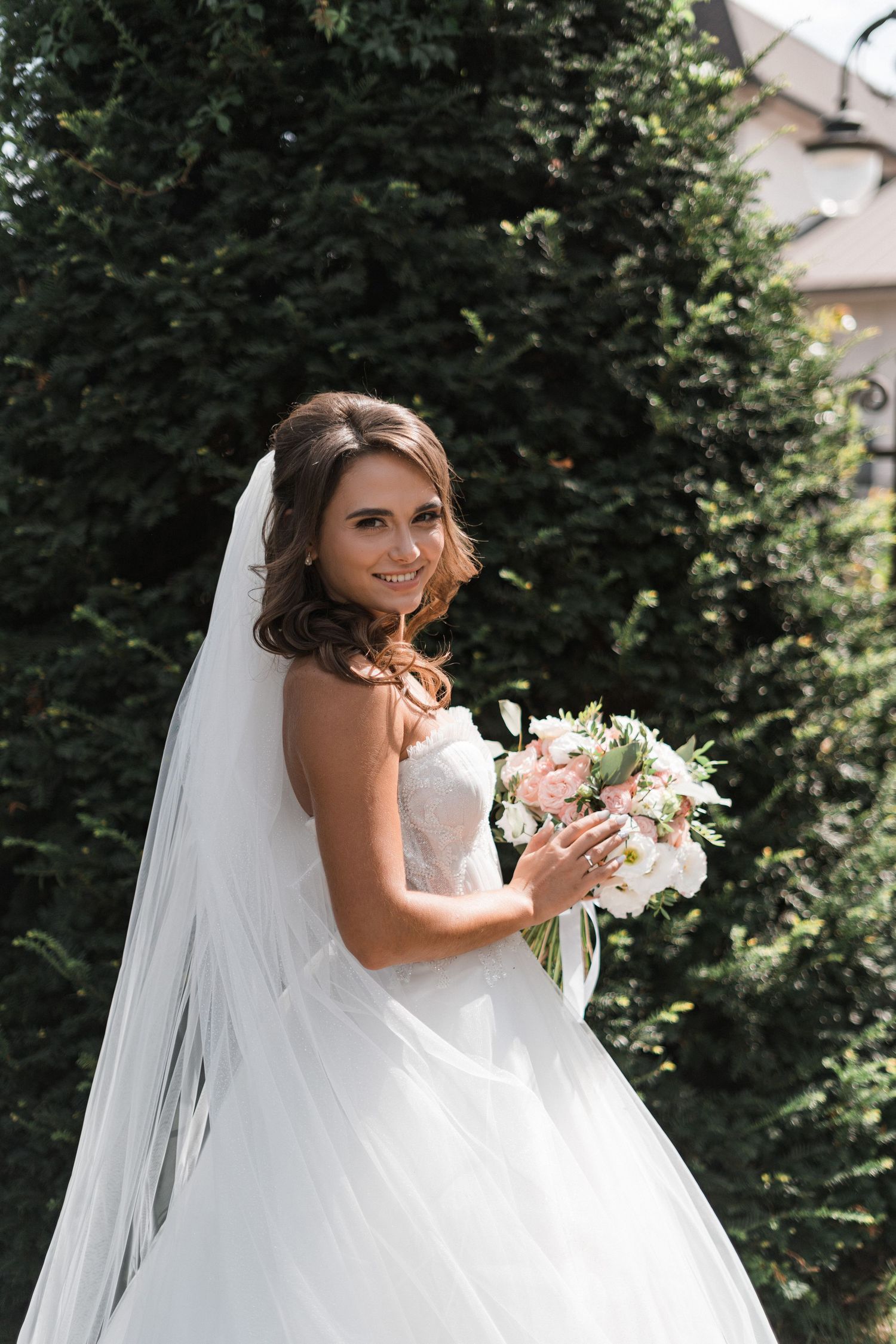 A bride in a white strapless wedding gown holds a soft pink and white bouquet against a backdrop of evergreen trees.