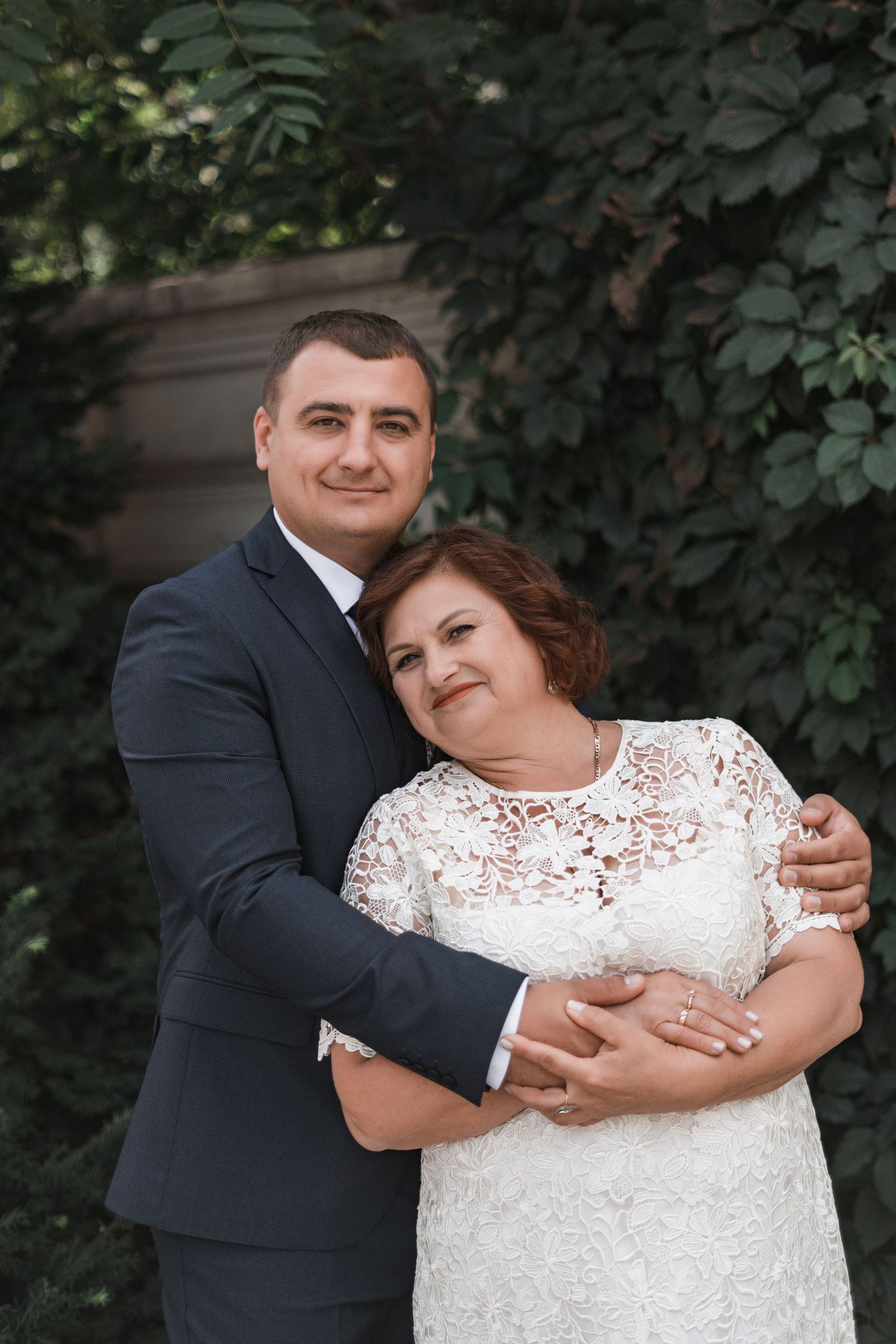 A couple embraces outdoors in wedding attire, with the bride wearing a white lace dress and groom in a dark suit.