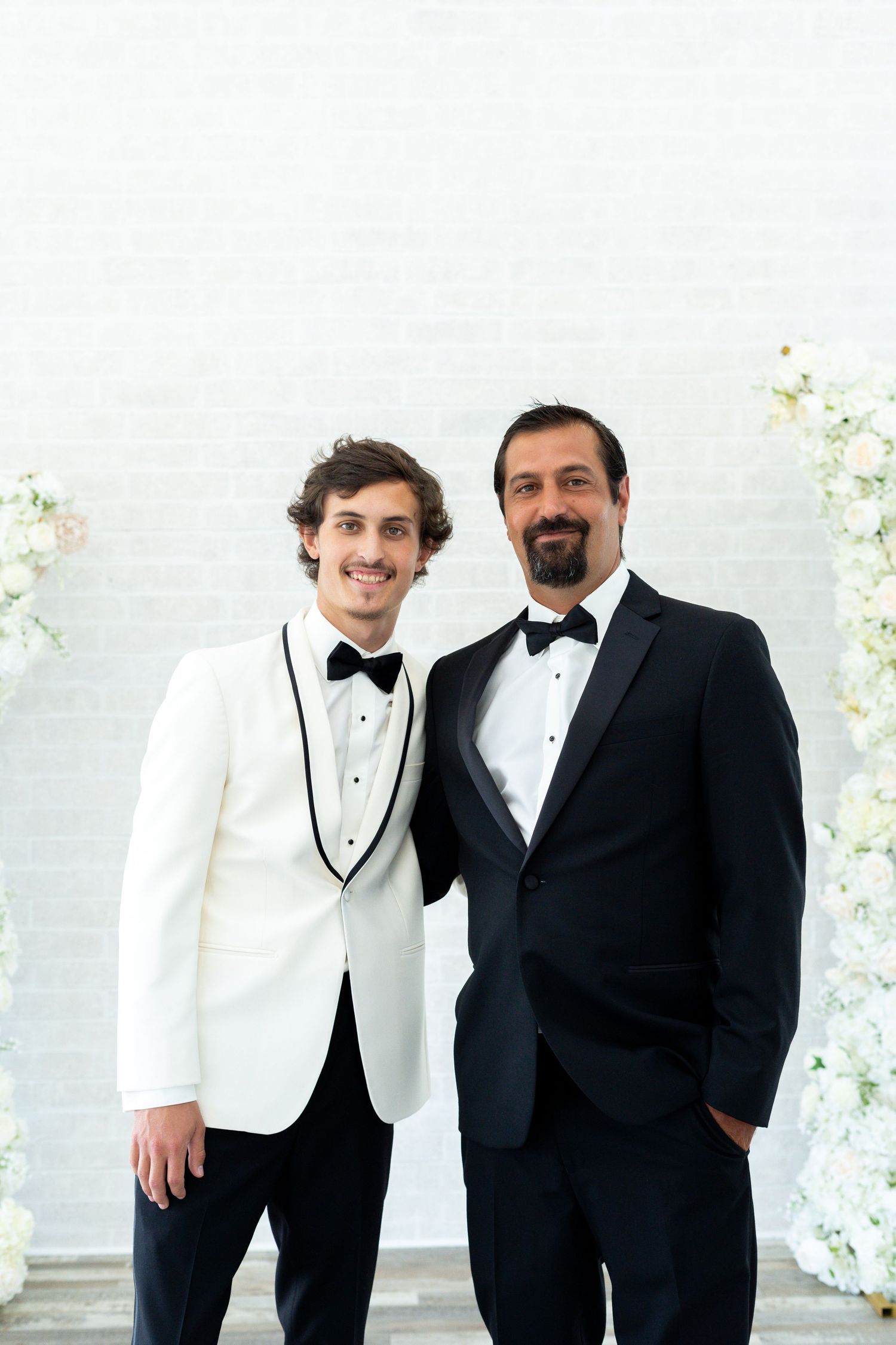 Two people in formal tuxedos pose together at an event with floral decorations in the background.