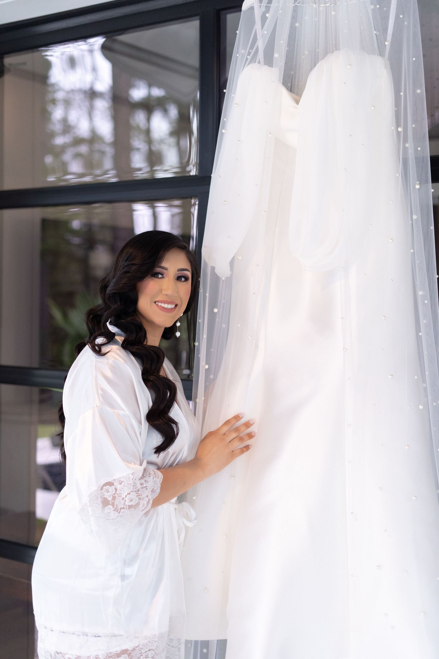 A bride in a white silk robe admires wedding dresses hanging on display in a bridal boutique.