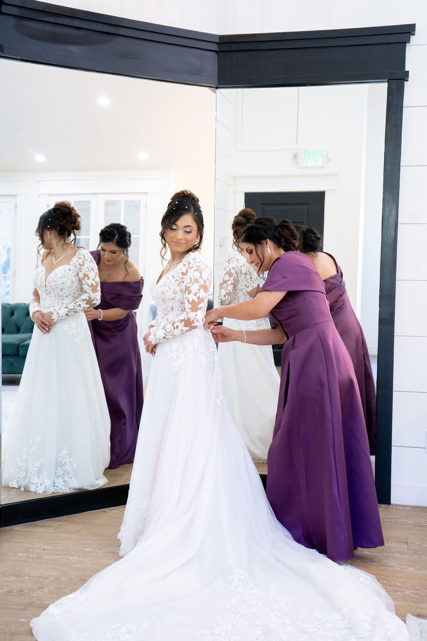 Bridesmaids in purple dresses help with final wedding dress adjustments in front of a mirror.
