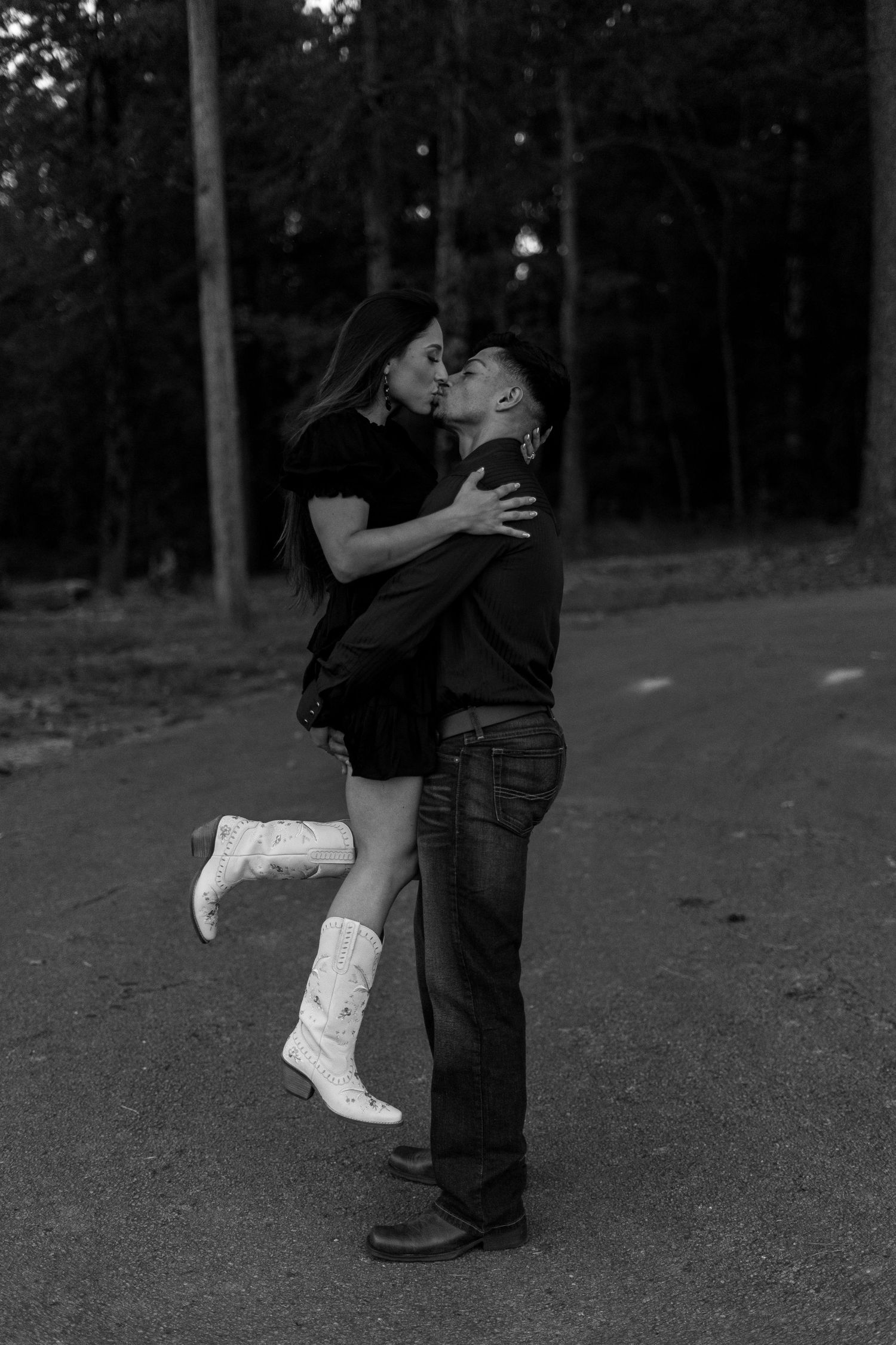 A couple in western wear shares romantic moments in black and white photography against a wooded backdrop.