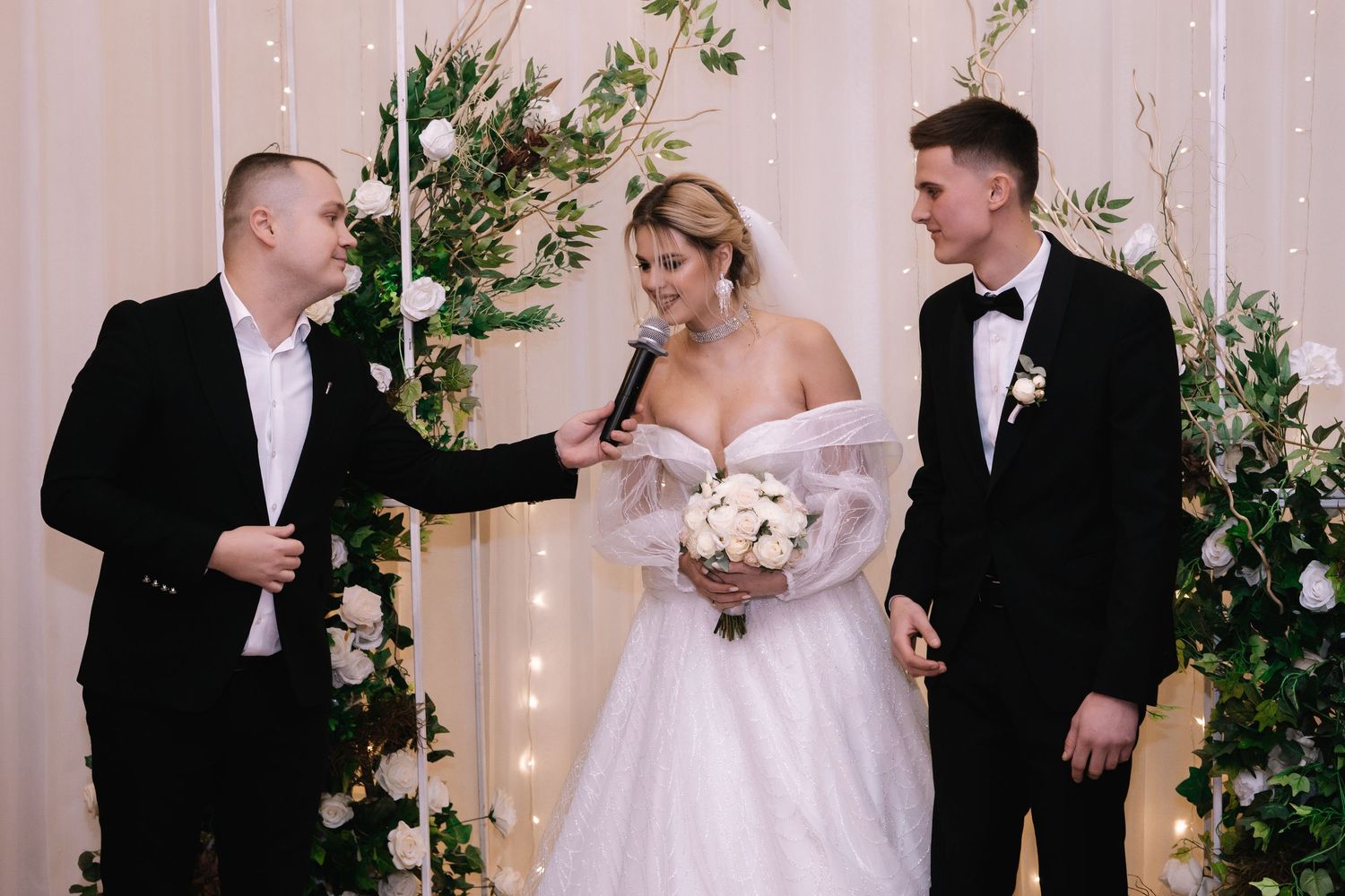 A wedding ceremony takes place under an elegant floral arch with twinkling lights and white decor.
