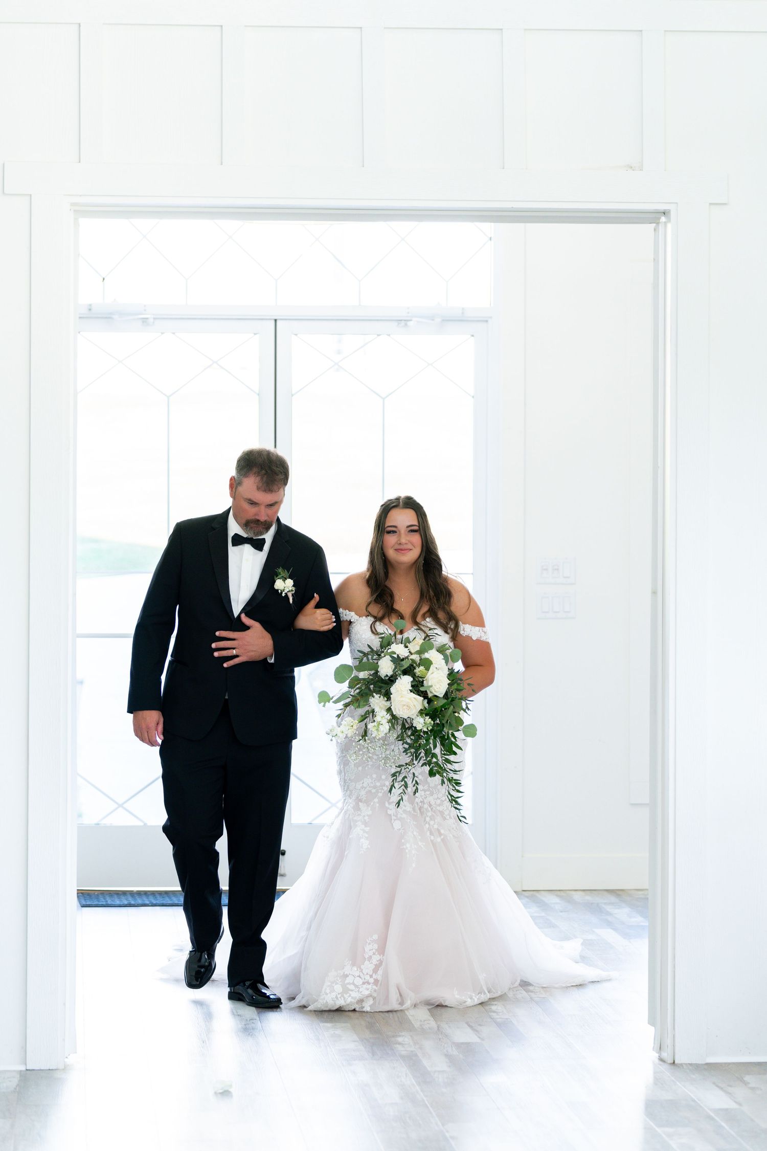 A wedding couple in white dress and black tuxedo walk through a bright modern venue with white walls.