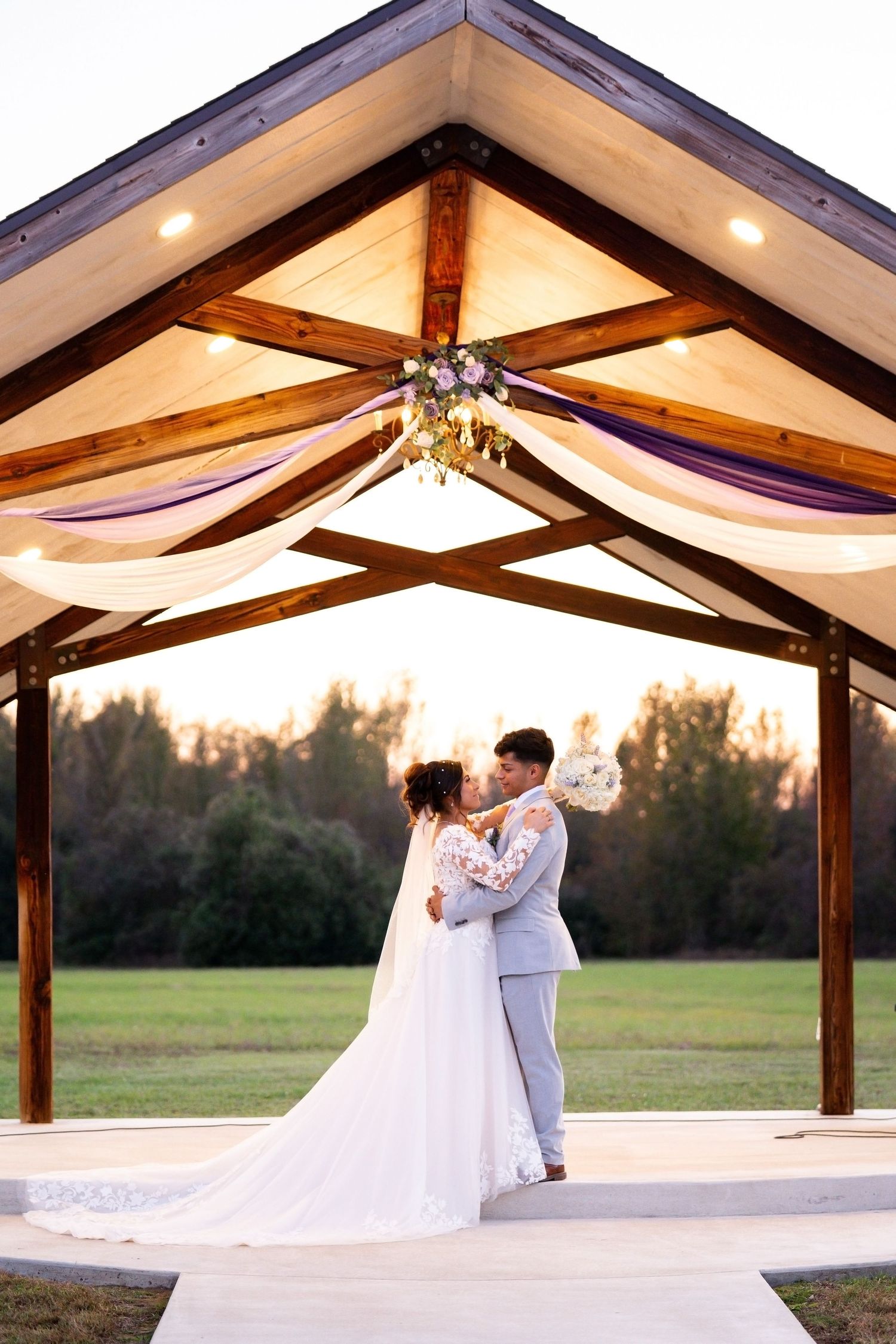 Wedding couple poses under wooden pavilion with draped fabric ceiling at sunset.