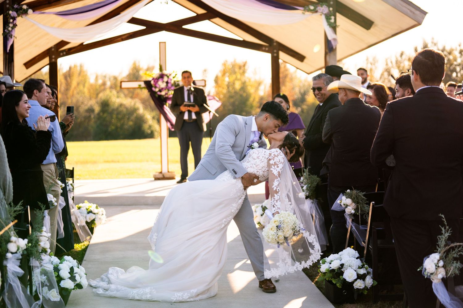 Wedding ceremony taking place under a rustic wooden beam venue with guests seated outdoors.
