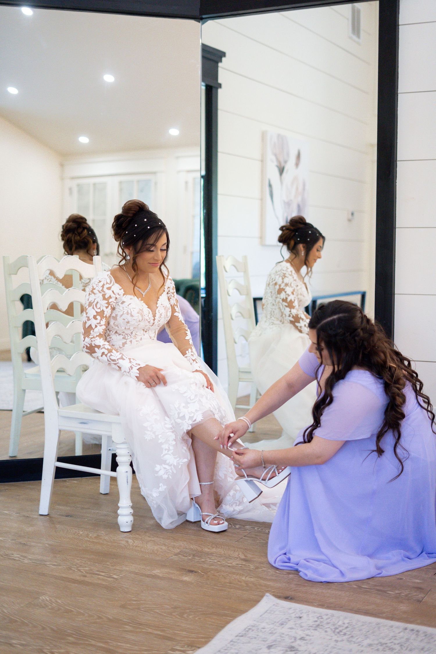 Wedding party members in purple and white dresses help with dress adjustments on hardwood floors.
