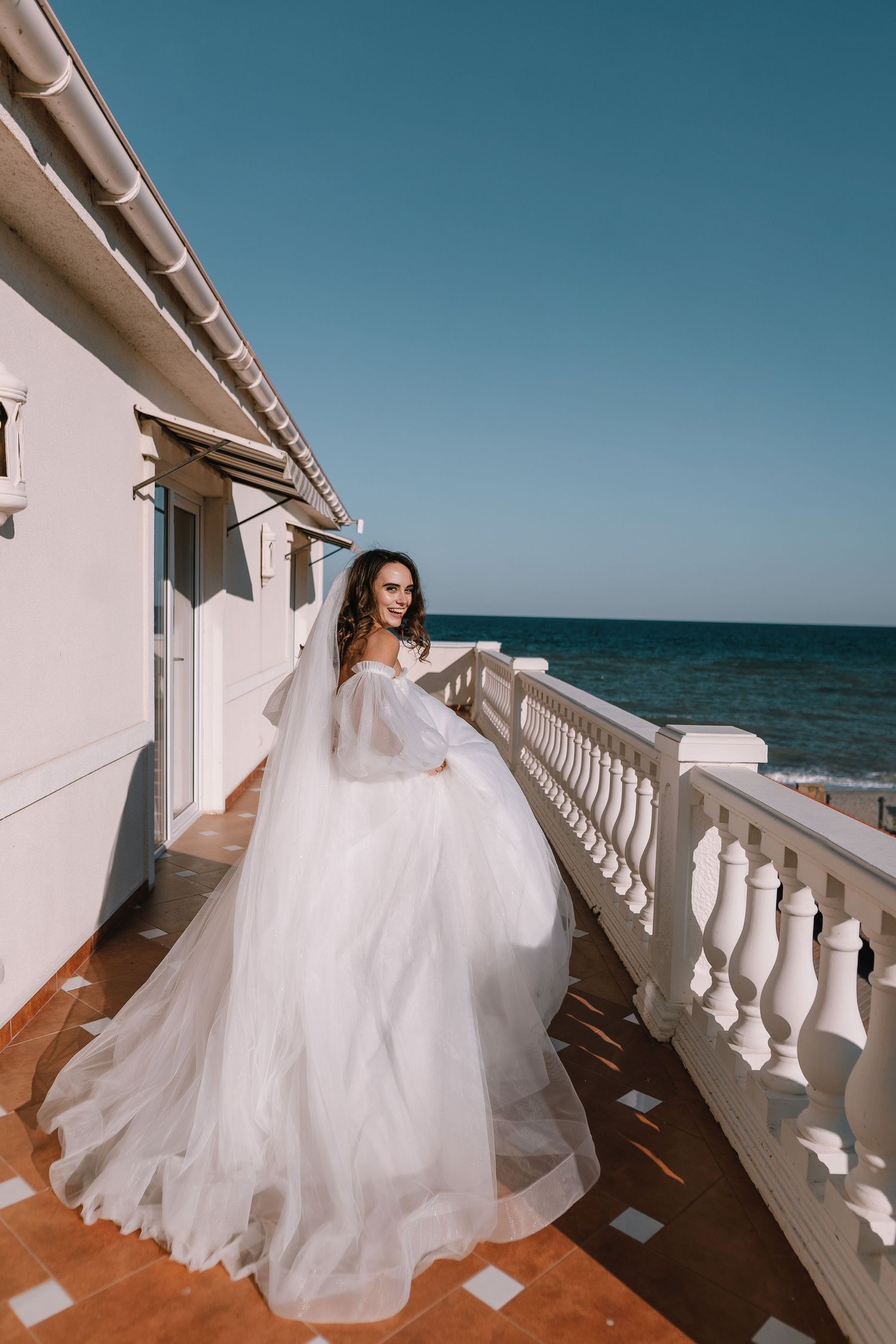 A flowing white wedding dress billows in the ocean breeze on a seaside balcony with white balustrades.