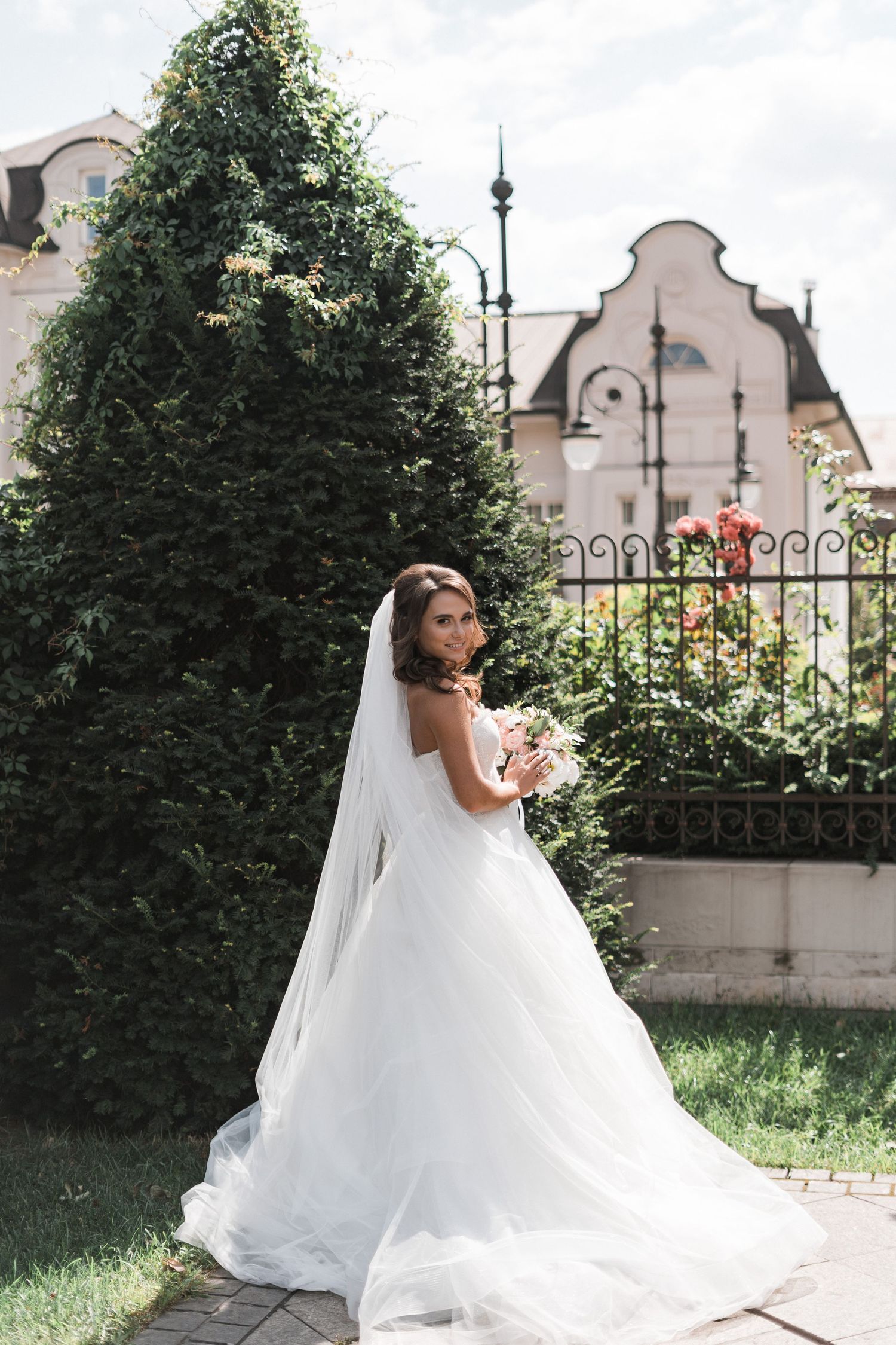 A bride in a flowing white wedding gown and veil poses gracefully in a garden setting with ornate architecture in the background.