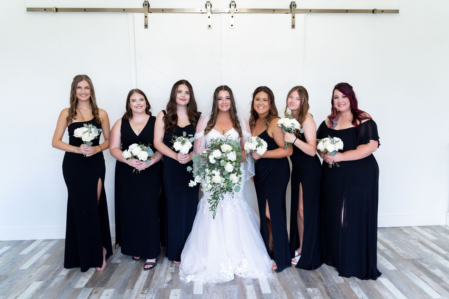 A group of bridesmaids in black dresses stand together with the bride in white holding bouquets at a wedding.
