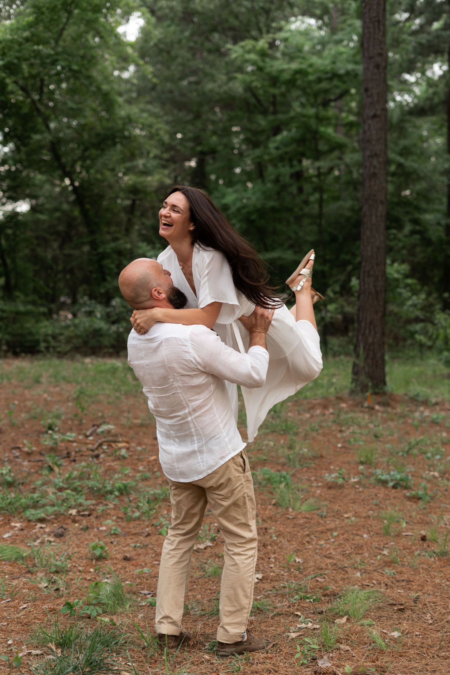 A joyful couple in white outfits shares a playful lifting moment in a pine forest.