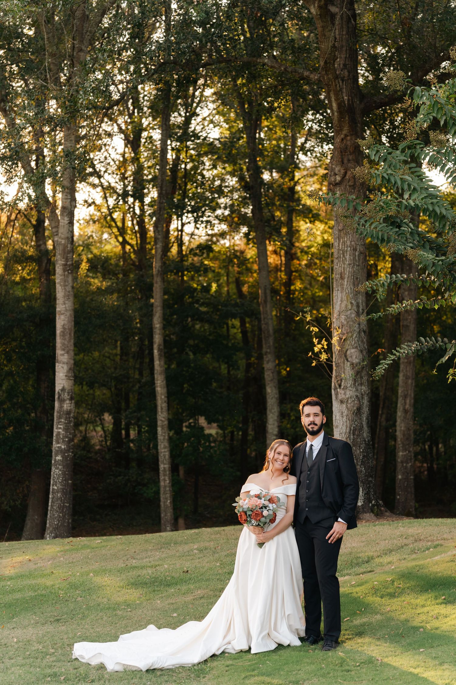 A wedding couple stands on a grassy slope at golden hour with tall trees creating a romantic forest backdrop.
