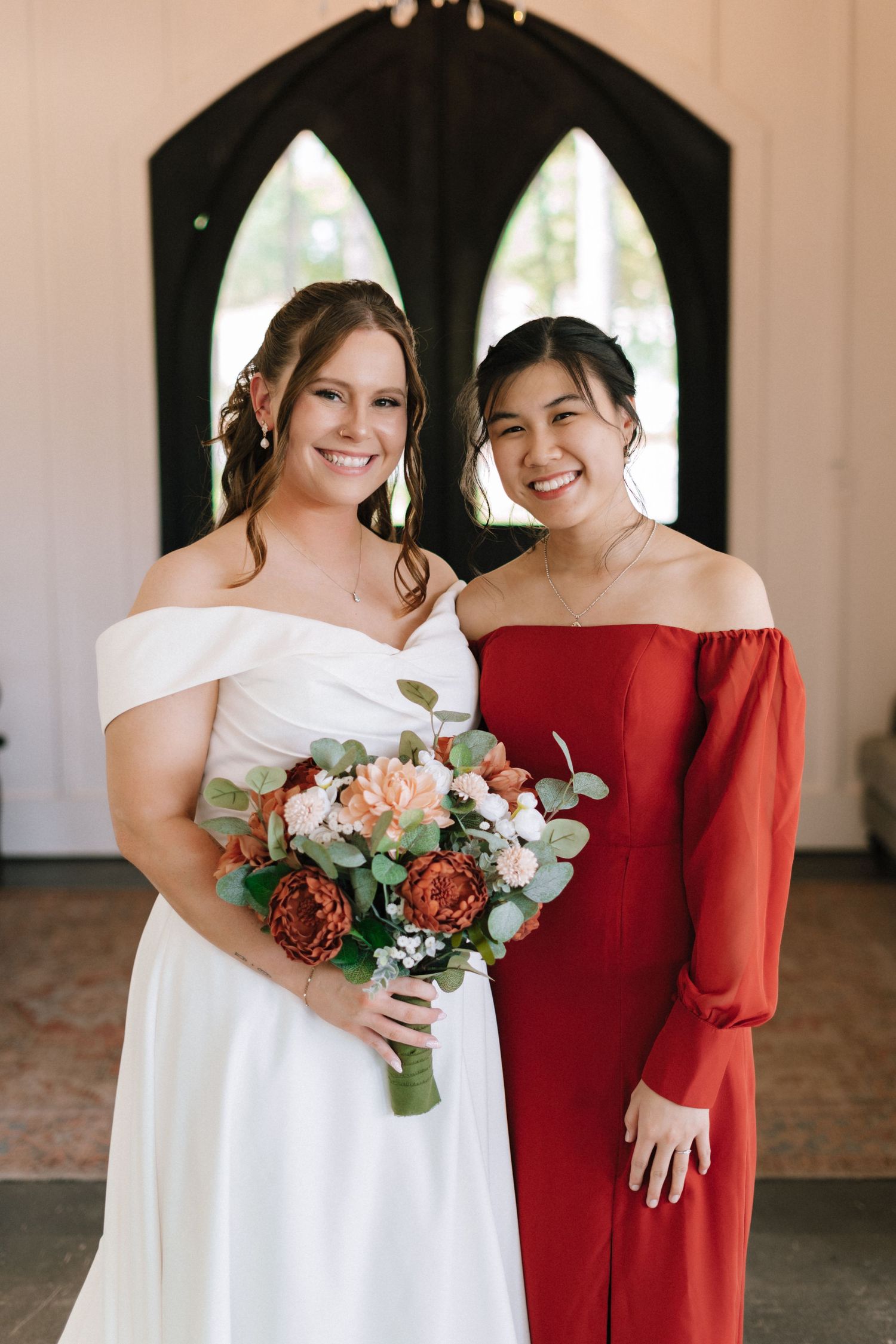 Two people pose together at a wedding, one wearing a white dress and one in a red dress holding floral bouquets.