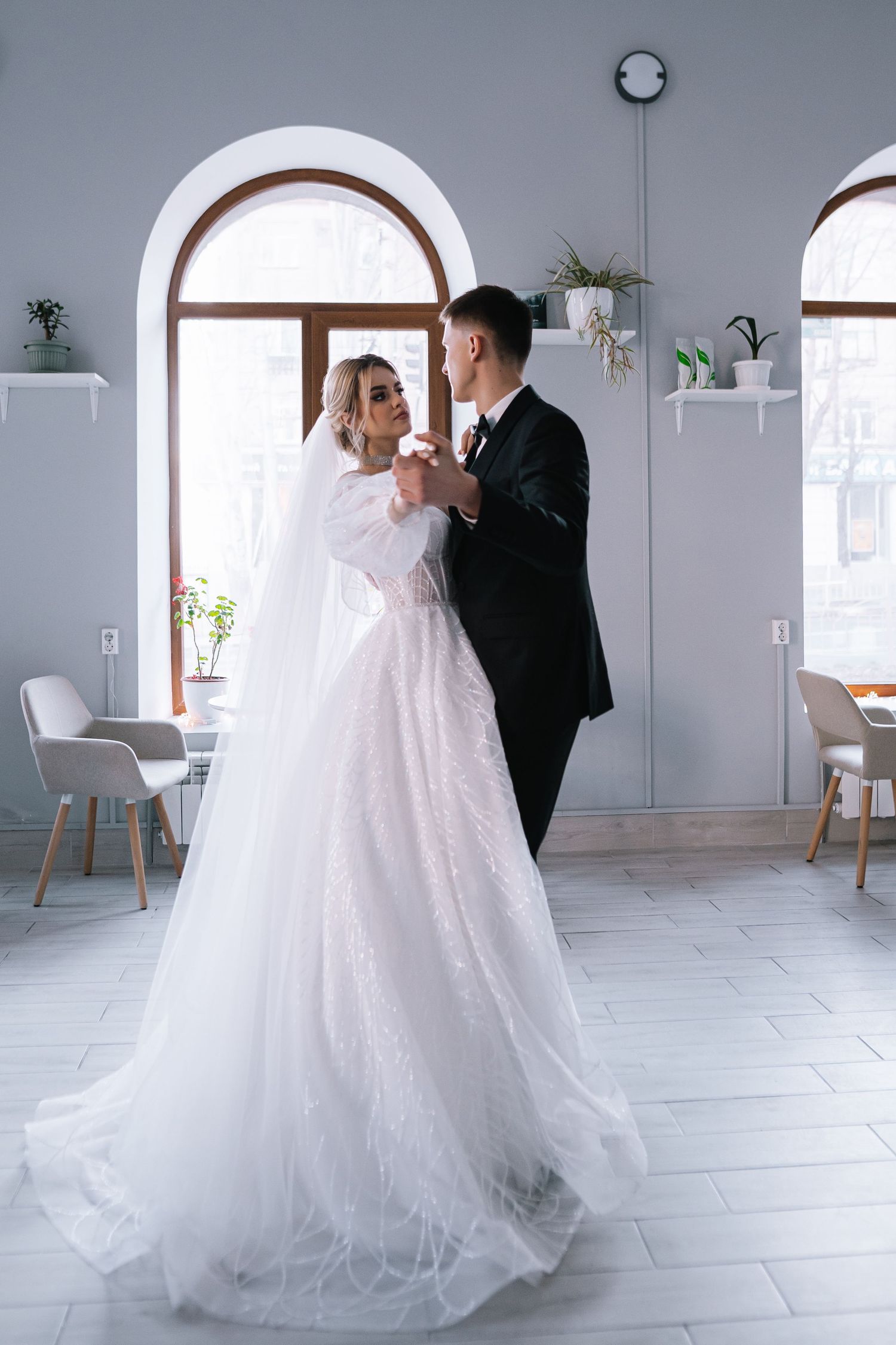A couple in wedding attire share an intimate dance moment in a bright, minimalist white room with arched windows.