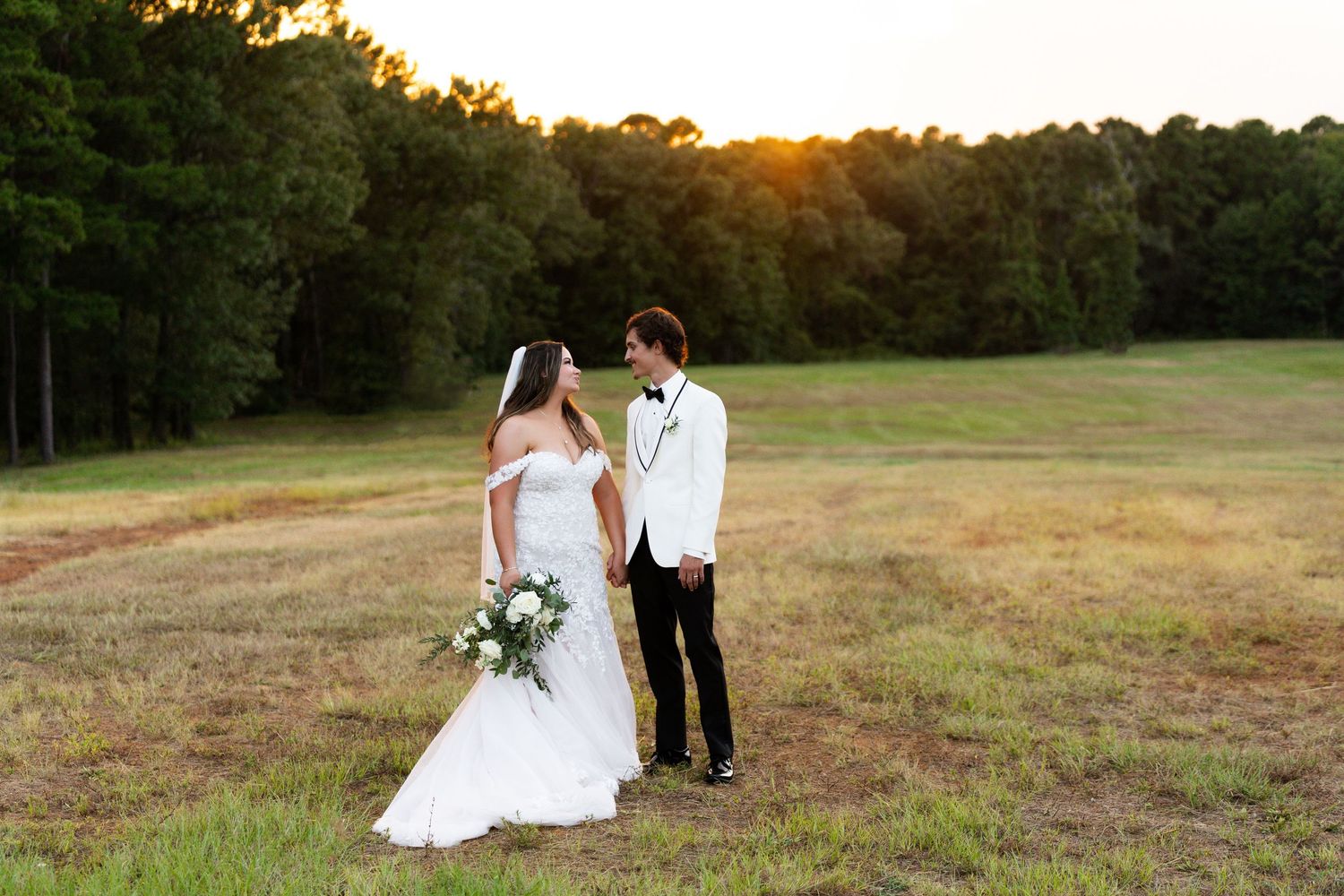 A couple in wedding attire stands in a golden field at sunset with trees in the background.