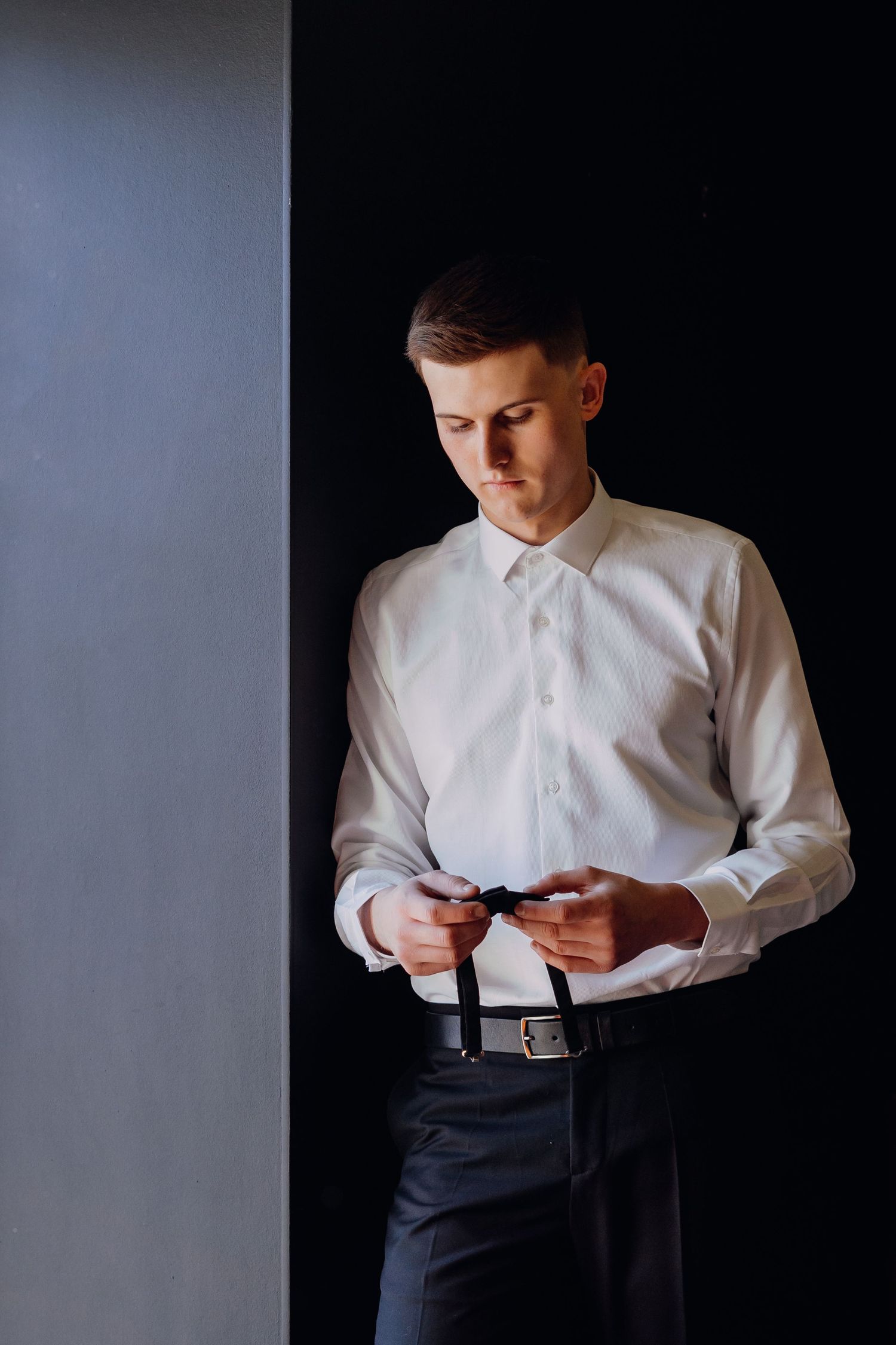 A person in a white dress shirt and dark pants buttons up their clothing against a dark background.