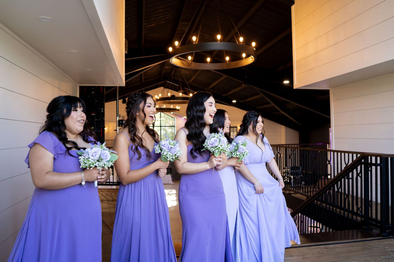 Wedding party members gather under rustic chandeliers in an elegant venue with white shiplap walls.