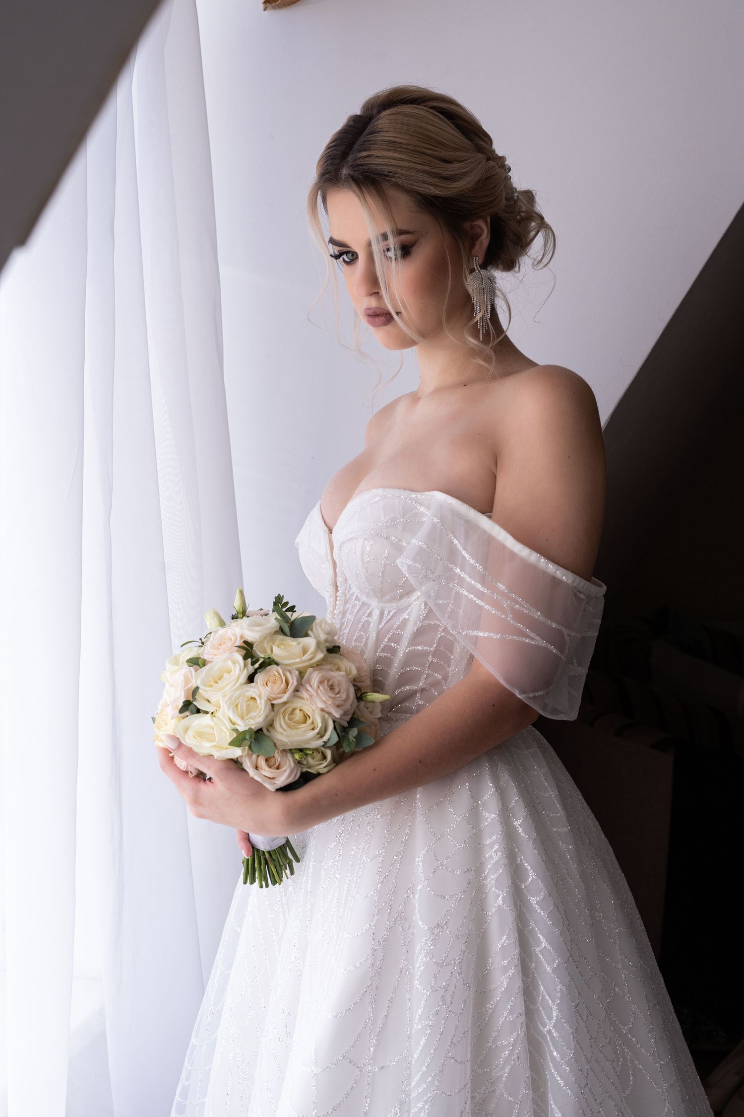A bride in an off-shoulder white wedding gown holds a delicate bouquet of cream roses against a light backdrop.