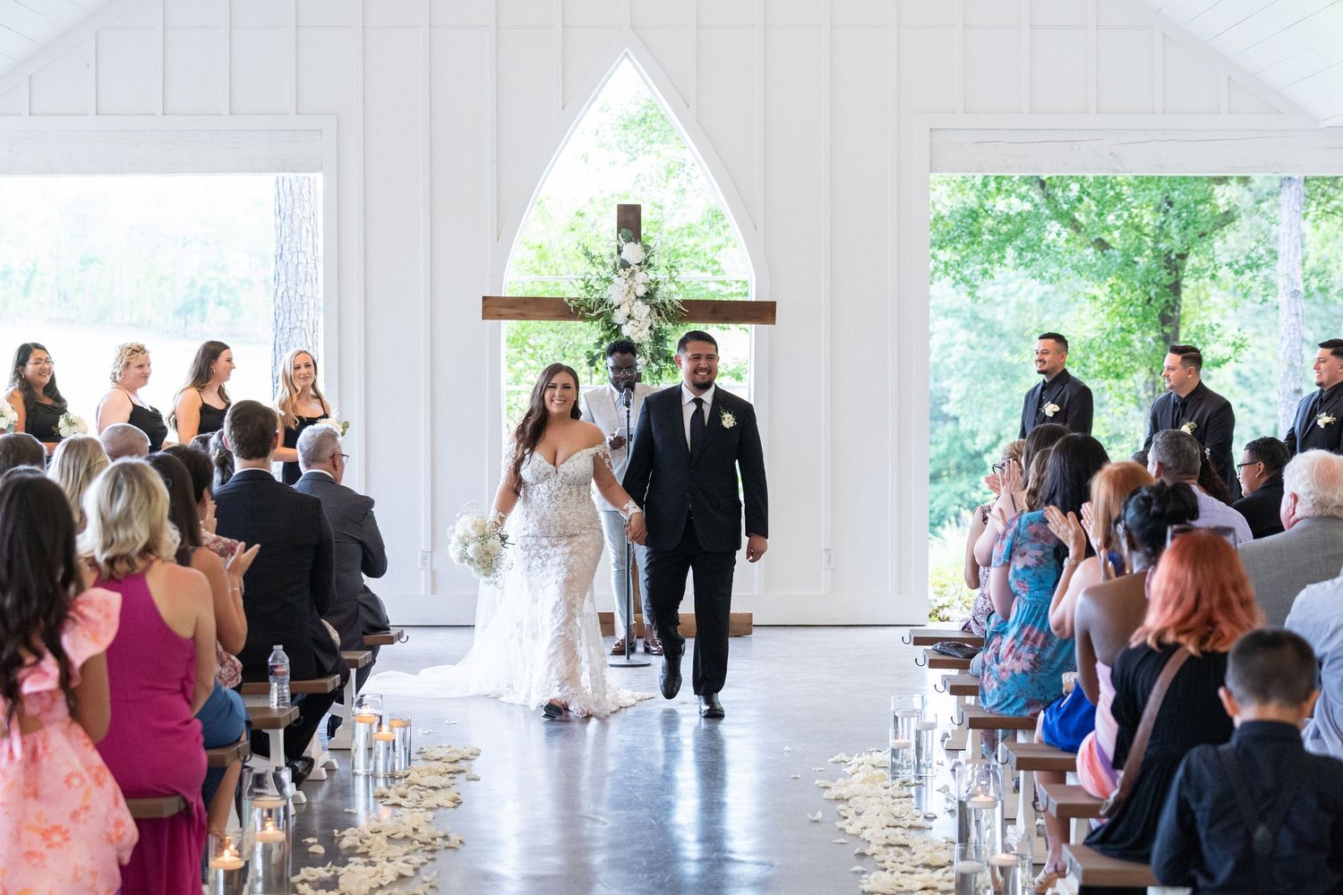 Wedding guests gather in a bright church interior with white walls and large windows during a ceremony.