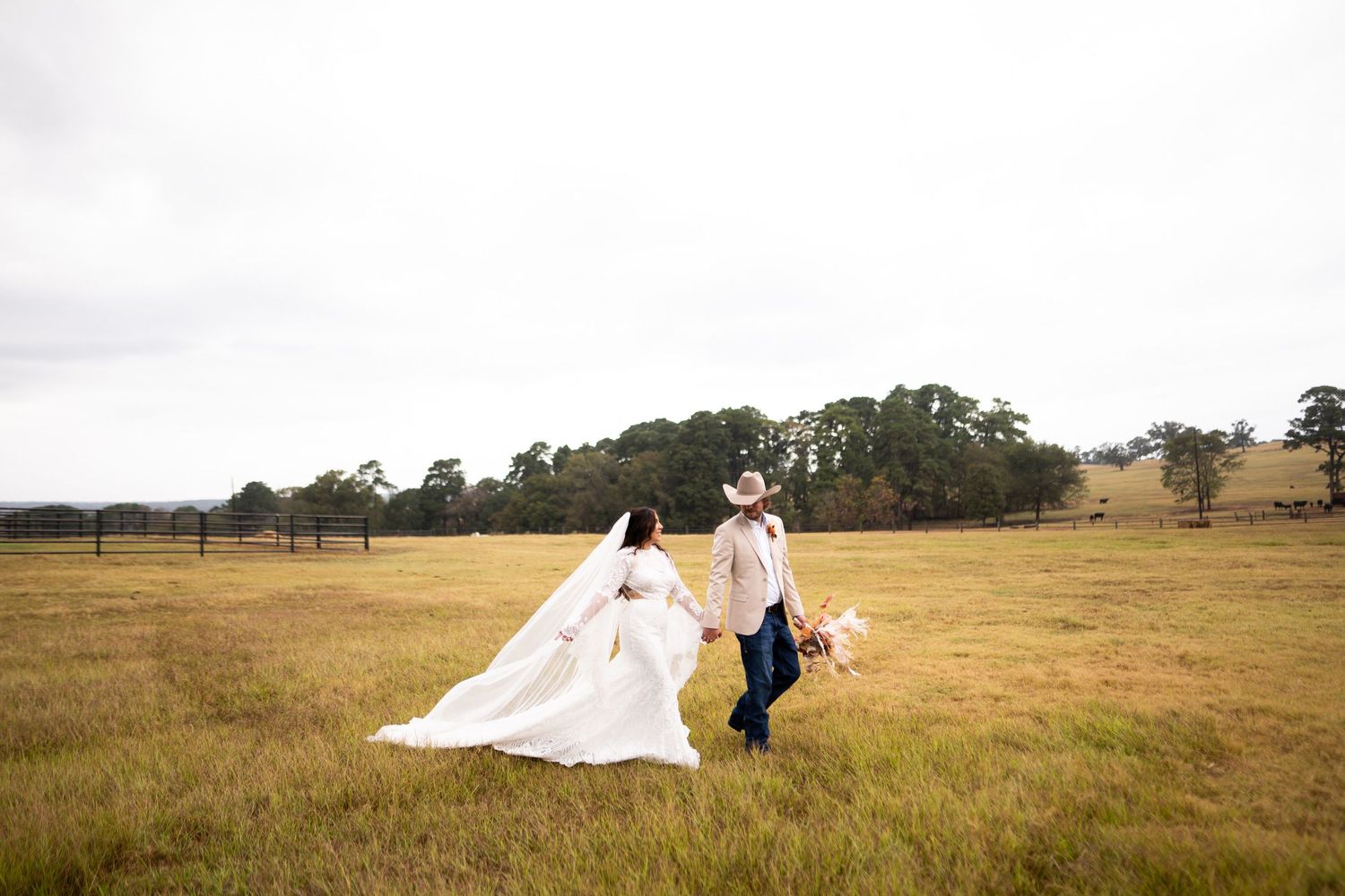 Wedding couple walking through golden grass field with flowing white dress trailing behind.