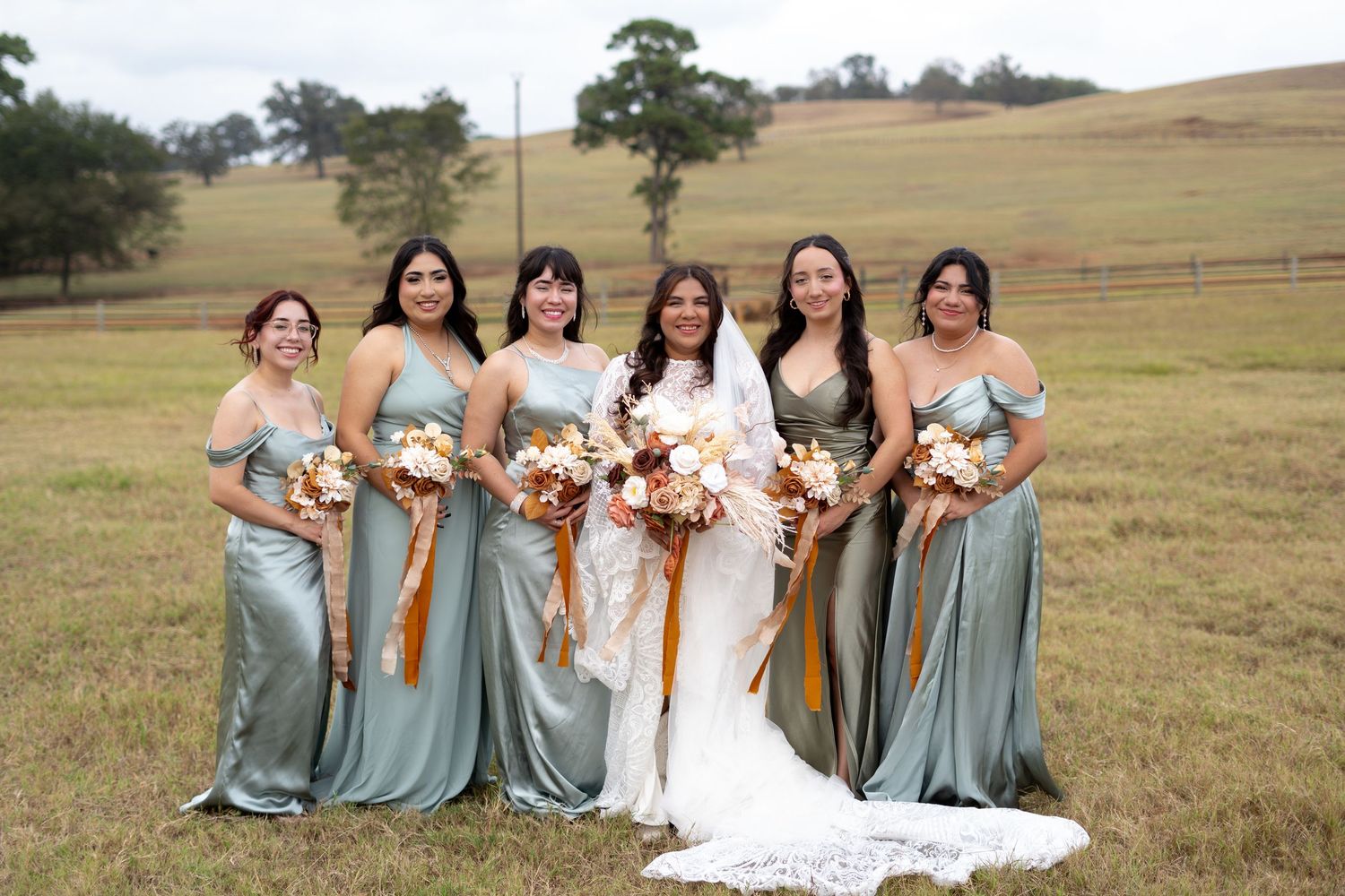 Bridesmaids in sage green dresses pose with bride in white gown holding autumn-colored bouquets in grassy field.