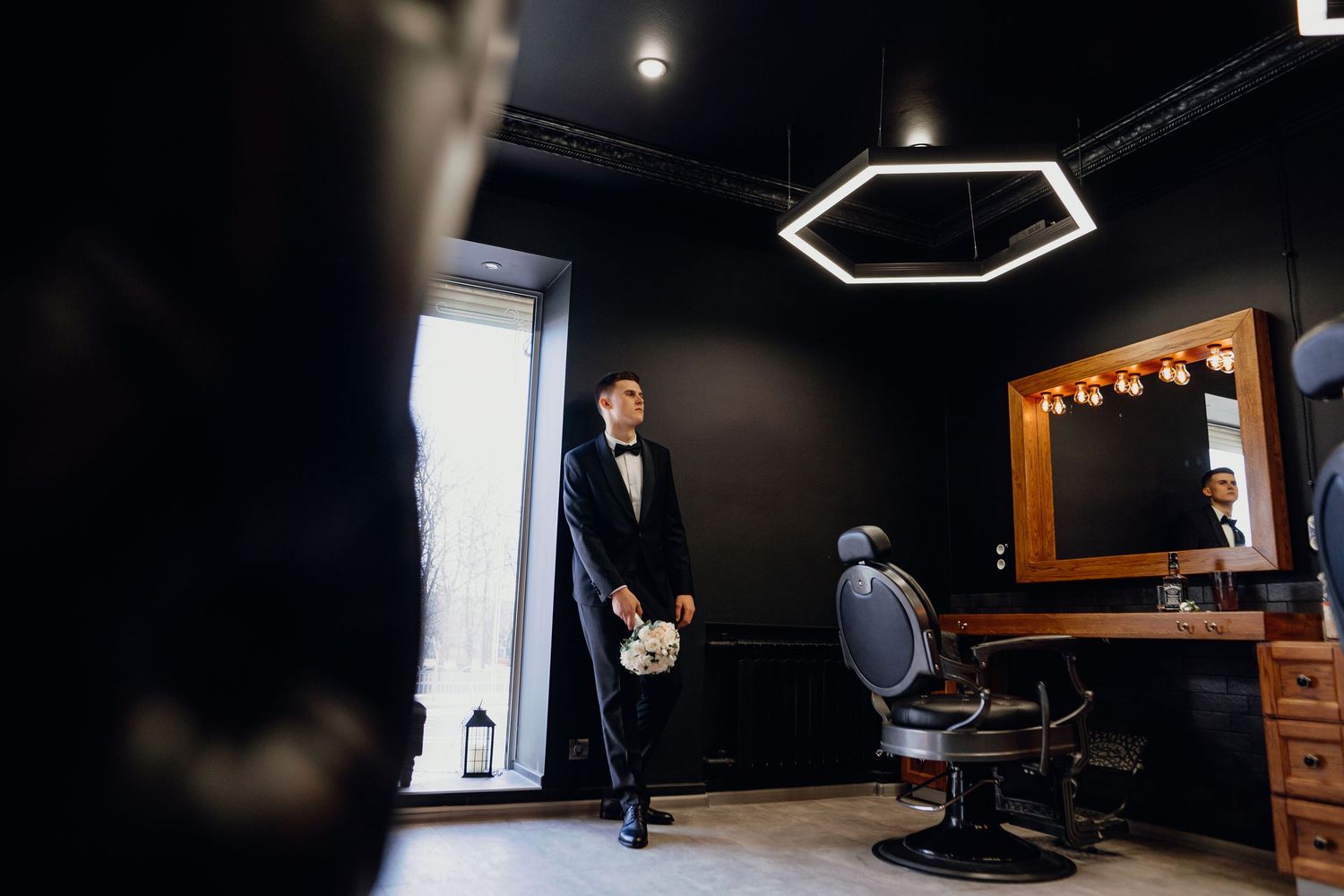 A modern barbershop interior with dark walls, hexagonal ceiling light, and mirror-lined station with leather chair.