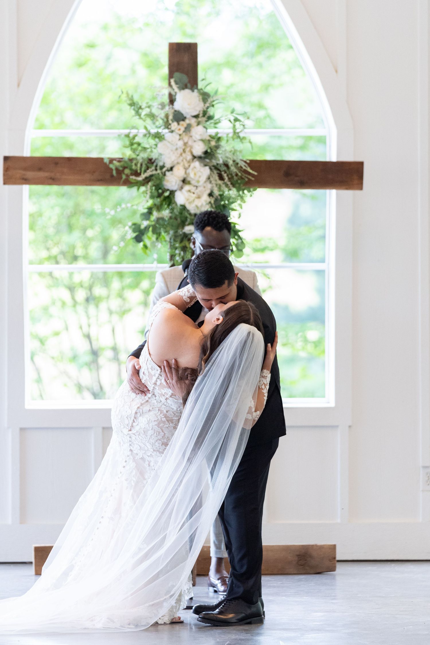 A couple shares a romantic dip kiss during their wedding ceremony beneath a wooden cross adorned with white floral arrangements.