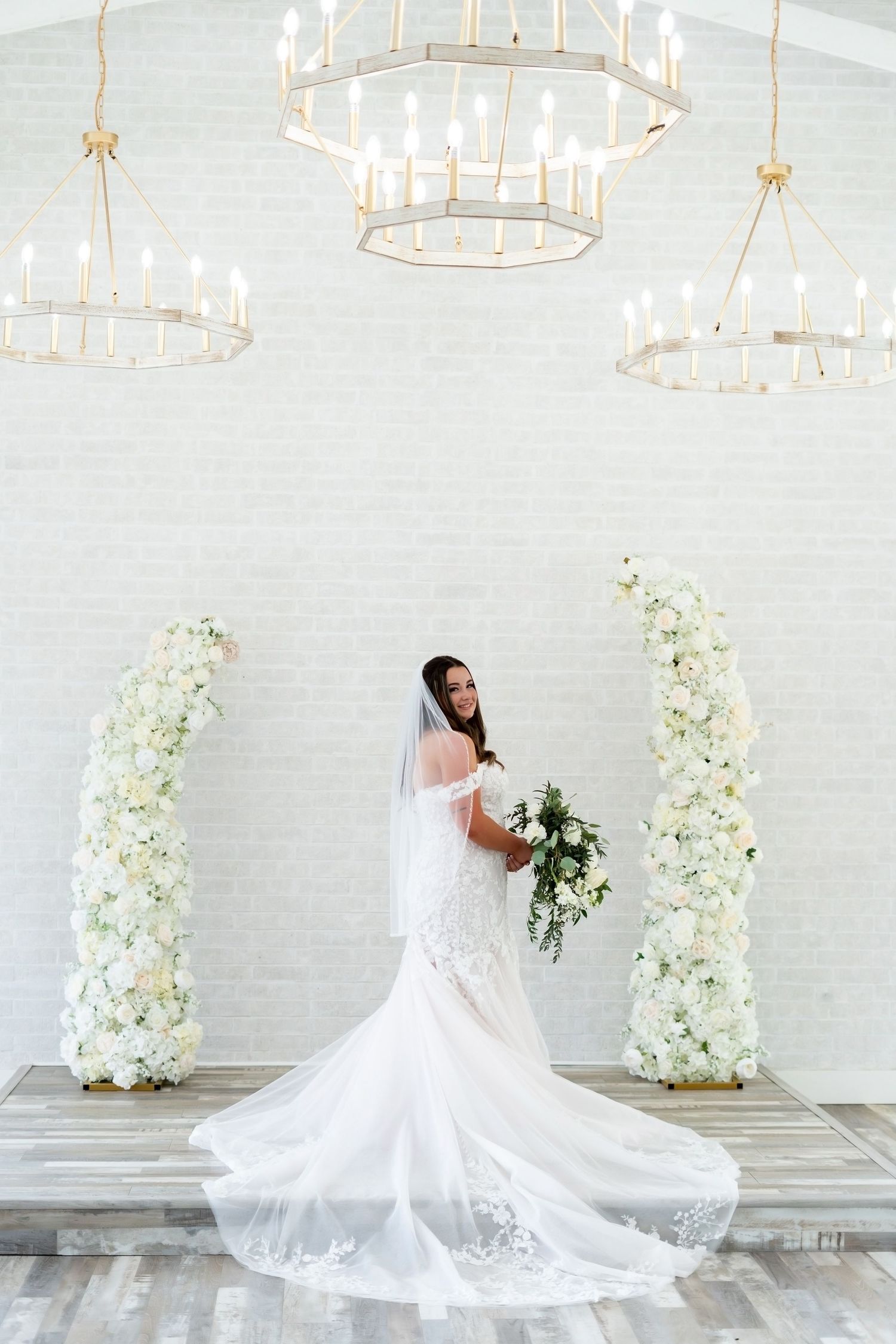 Elegant wedding scene with flowing white gown against white floral columns and illuminated chandeliers above.