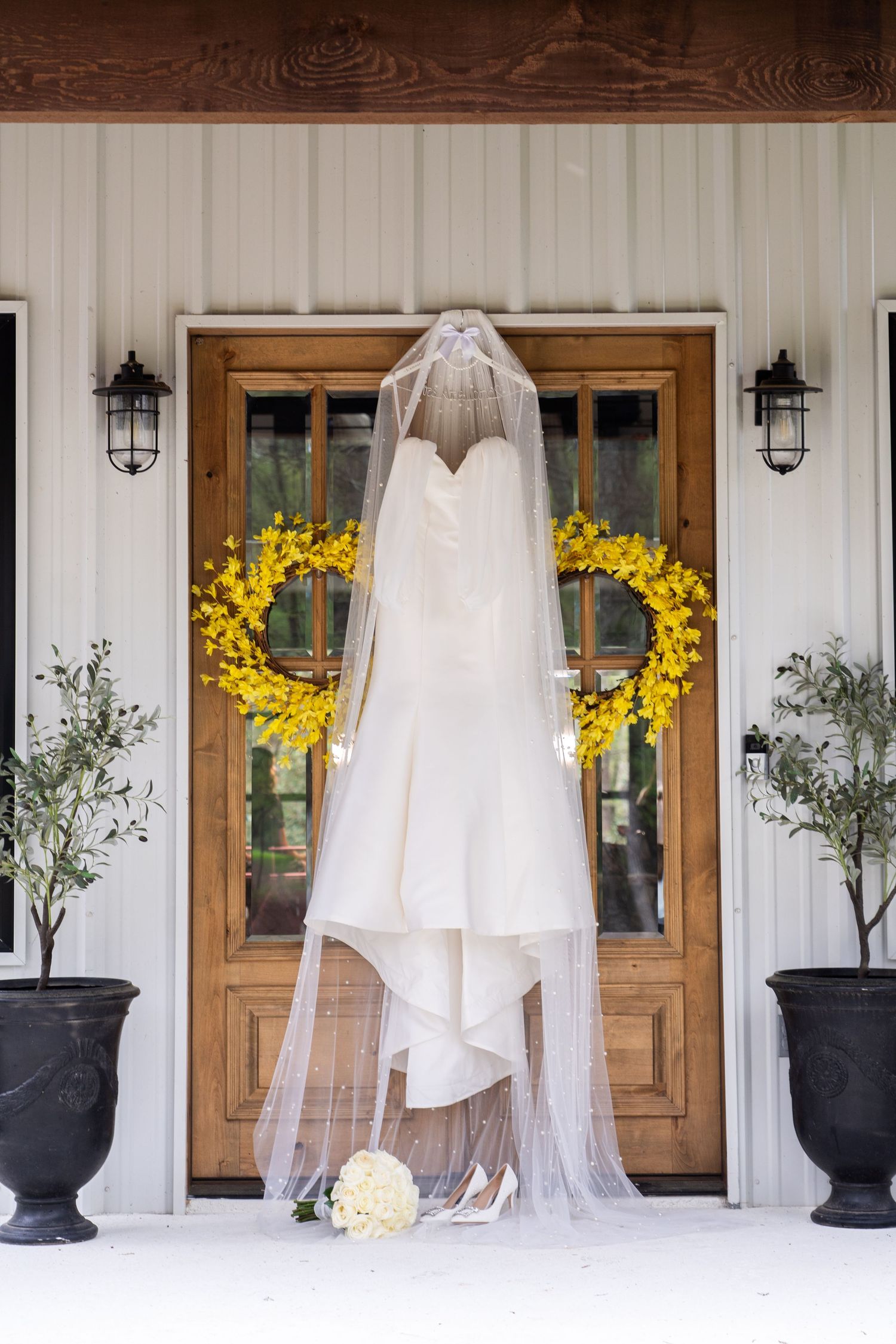Elegant white wedding dress hangs in front of rustic wooden doors with yellow floral arrangements and black planters.