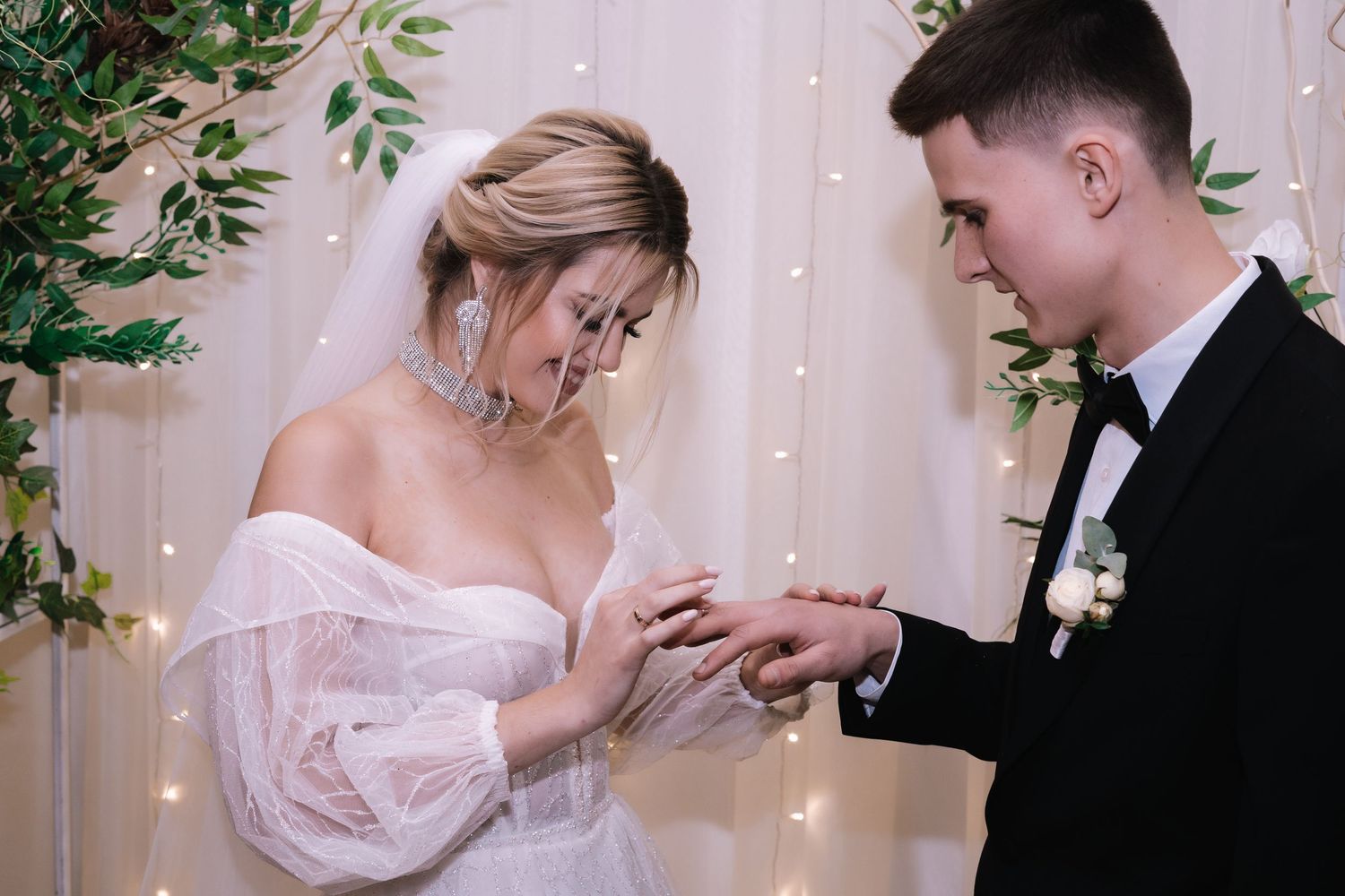 A couple exchanges rings during an elegant wedding ceremony with twinkling lights and greenery decorations.