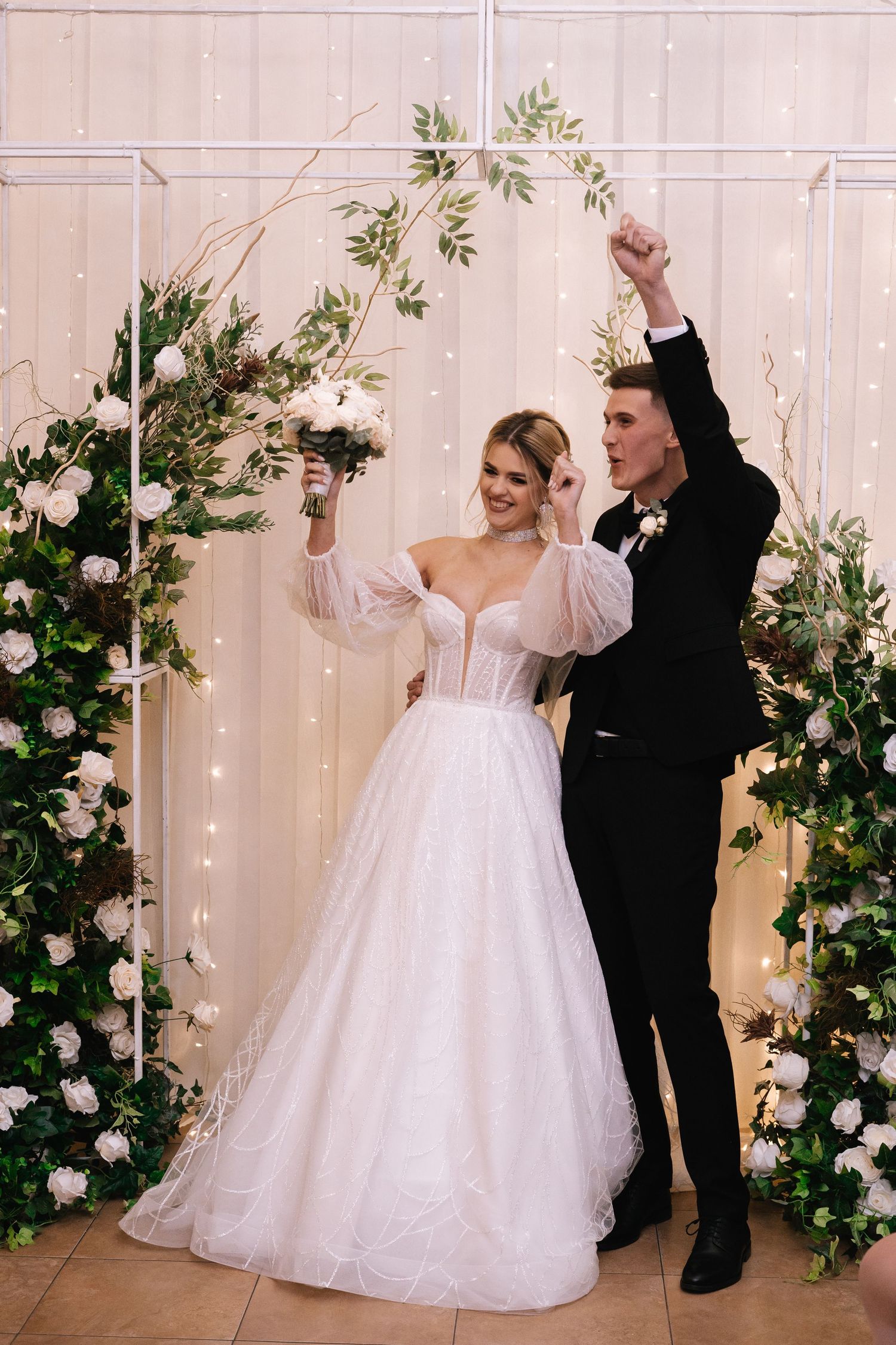 A bride in a white off-shoulder gown and groom in black tuxedo celebrate under a floral arch with twinkle lights.
