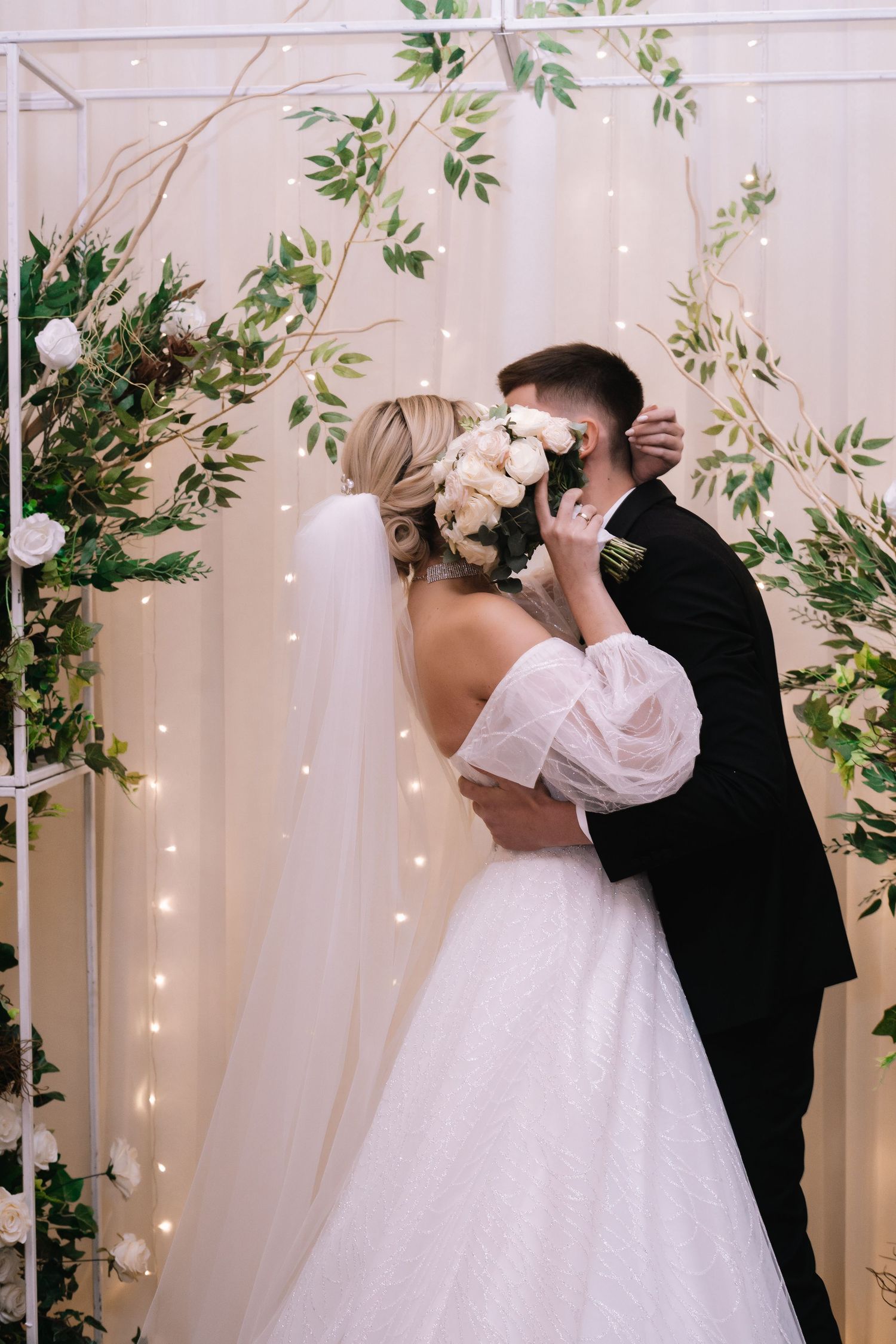A romantic wedding couple shares their first kiss as newlyweds under a floral arch with twinkling lights.