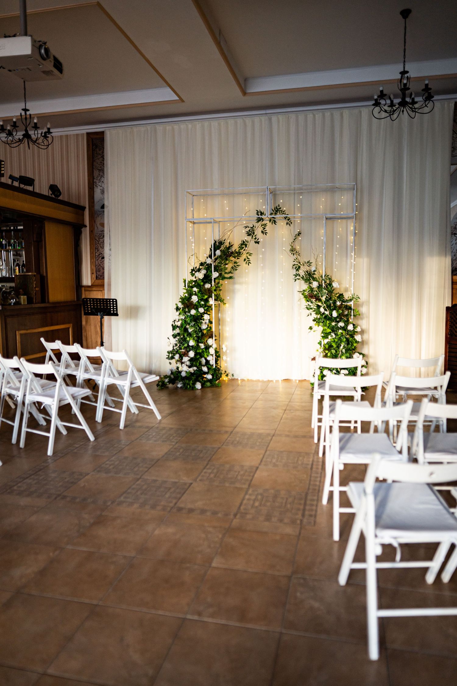 Indoor wedding ceremony space with white folding chairs and greenery decorations against white curtained backdrop.