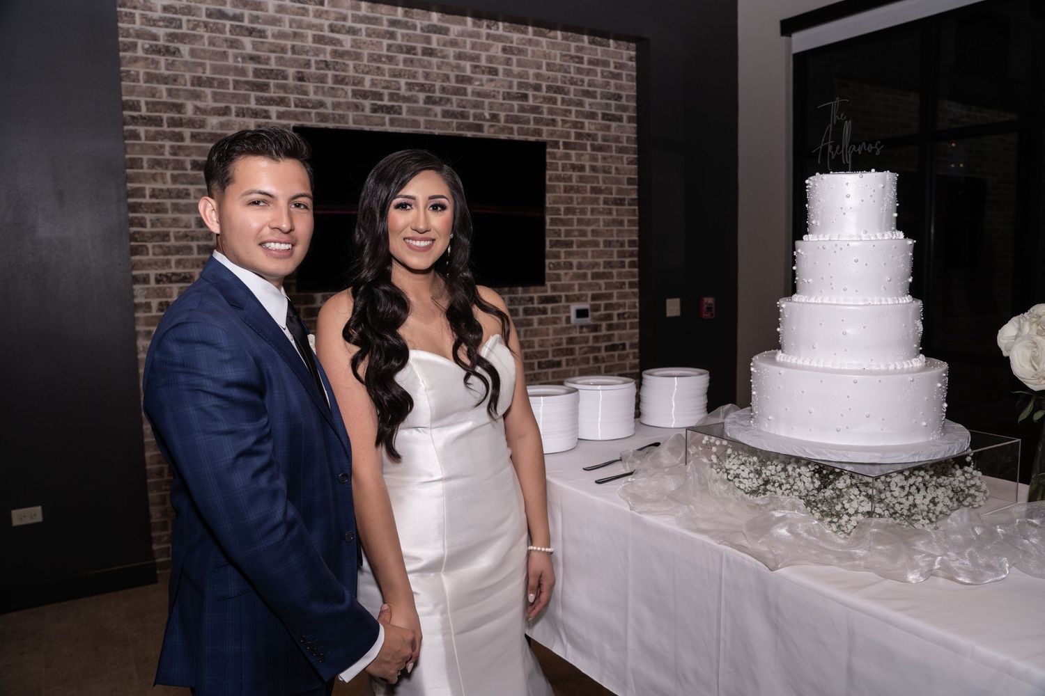 A couple stands next to their elegant four-tier white wedding cake at their reception with a brick wall backdrop.