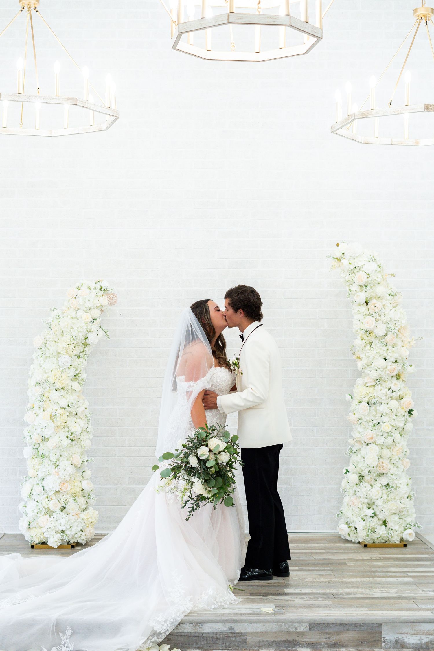 A romantic wedding scene with a floral arch backdrop and flowing white veil against a light colored wall.