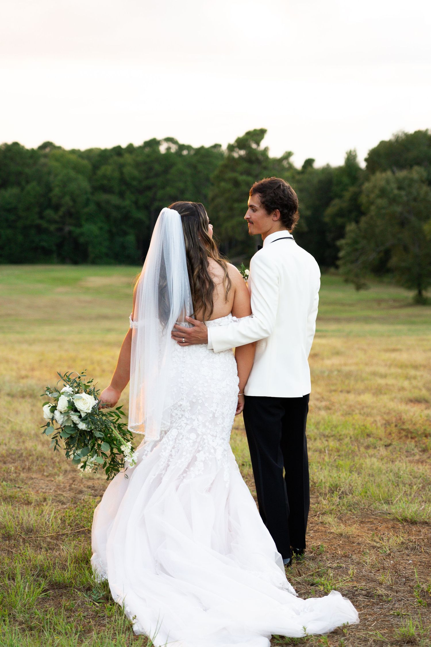 A romantic wedding couple walks together across a grassy field at sunset in matching white attire.
