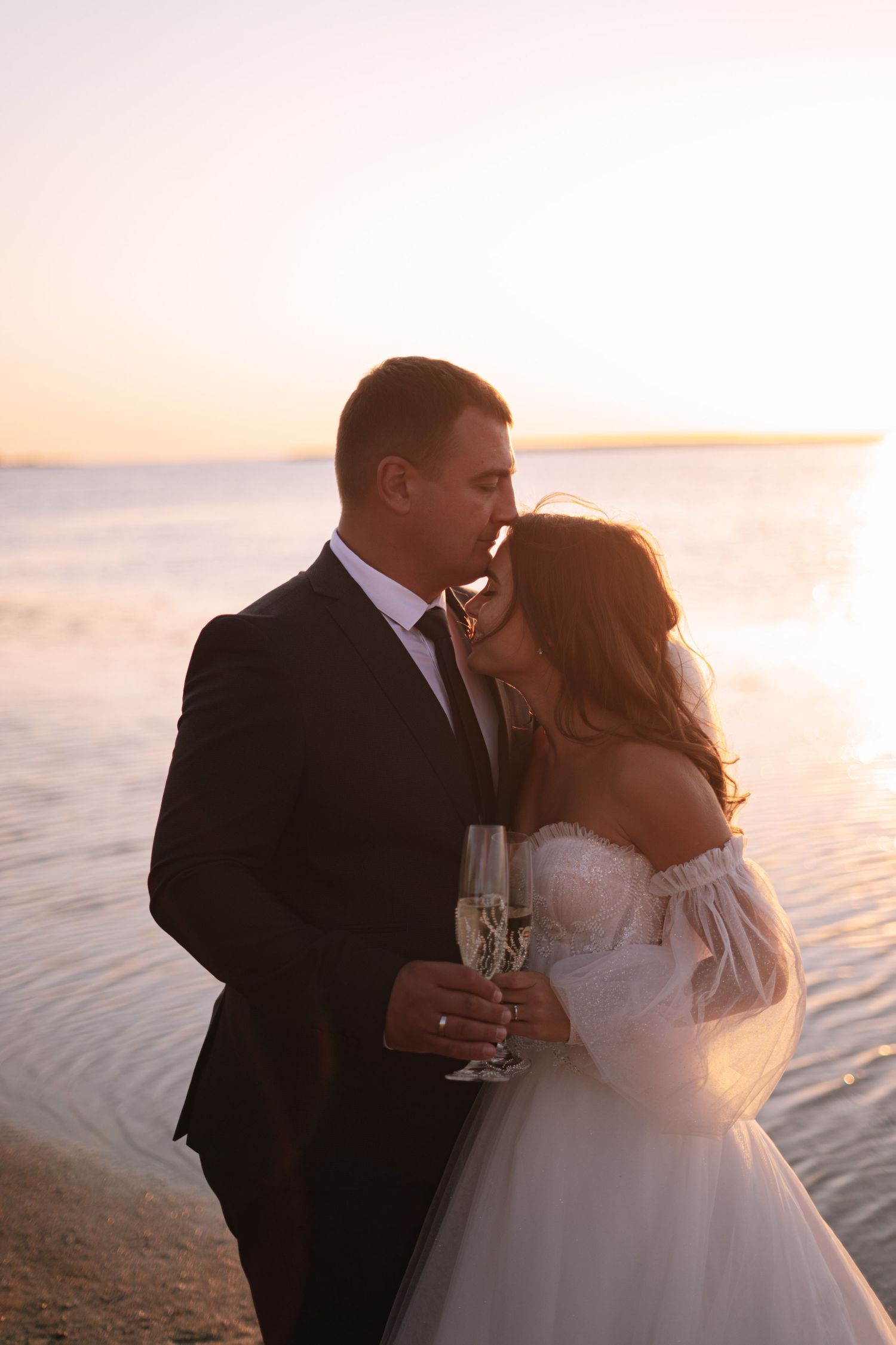 Romantic couple shares a tender kiss at sunset on the beach while holding champagne glasses in their wedding attire.