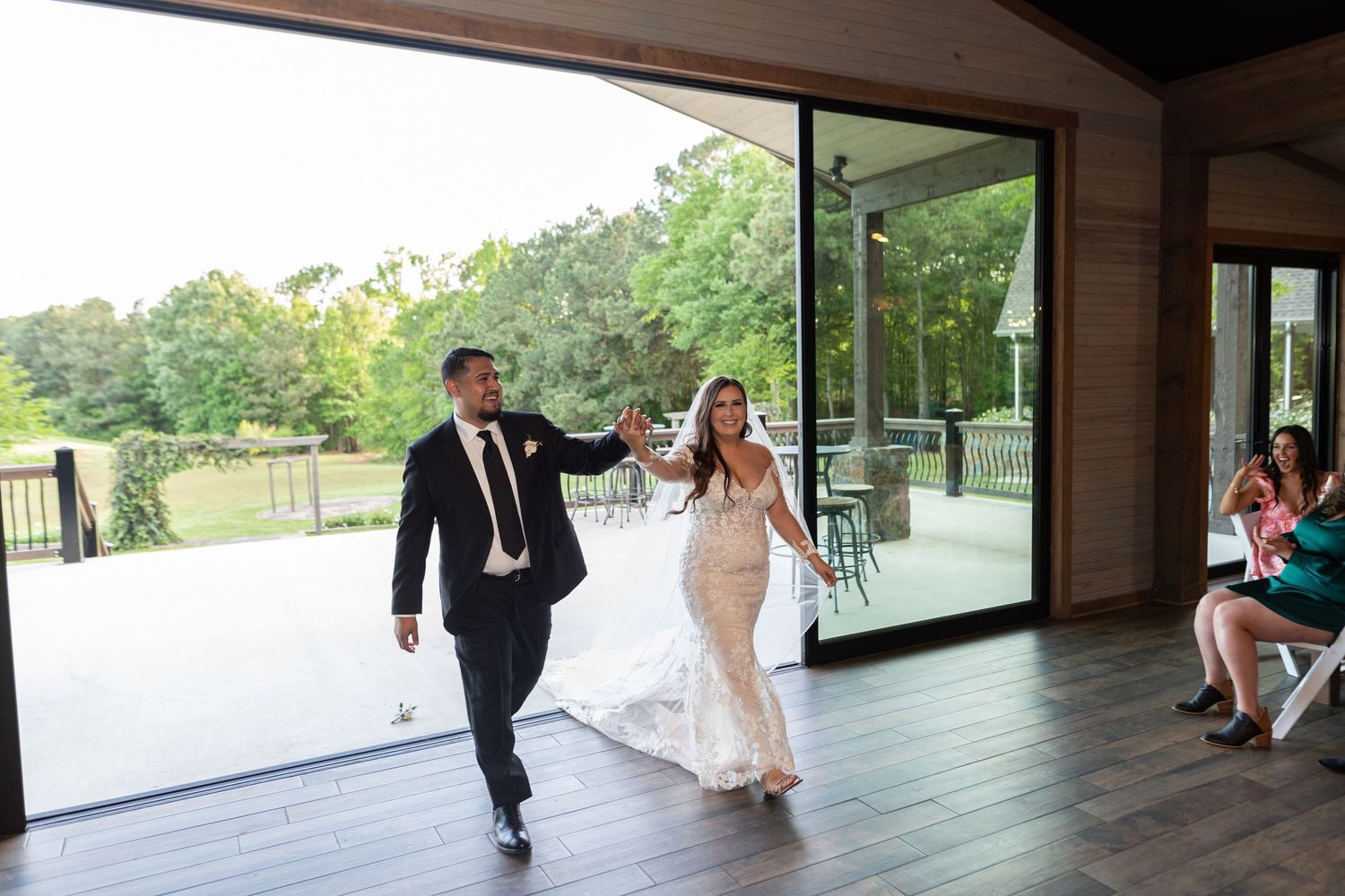 A couple in formal wedding attire joyfully enter their reception venue on a covered outdoor deck.