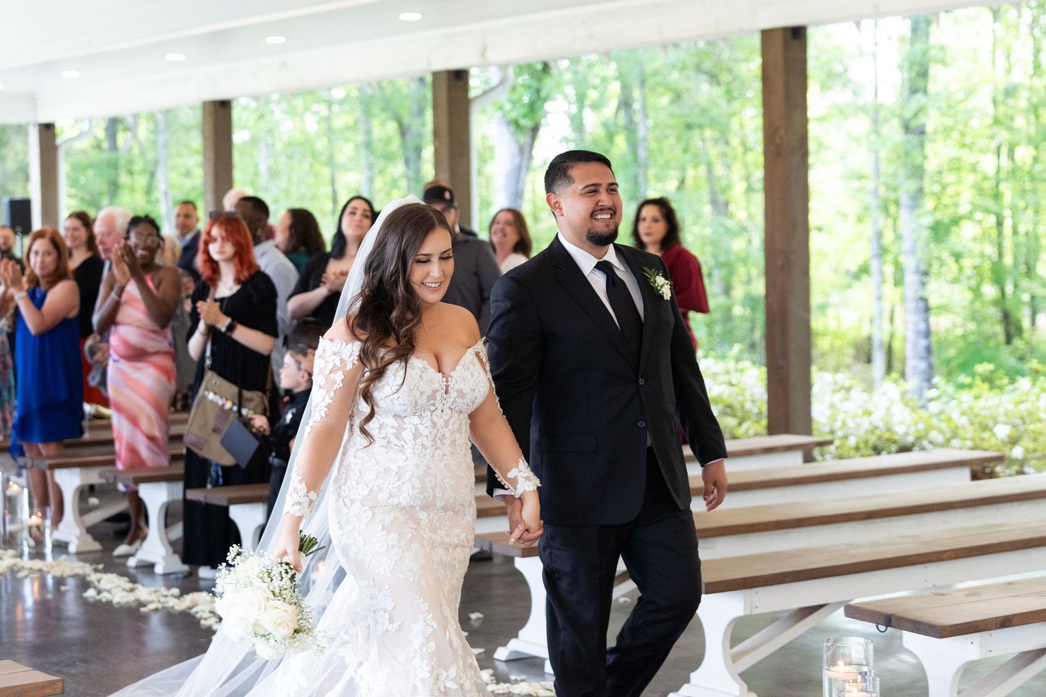 Newlyweds walk down the aisle at an outdoor covered pavilion venue surrounded by forest and wedding guests.