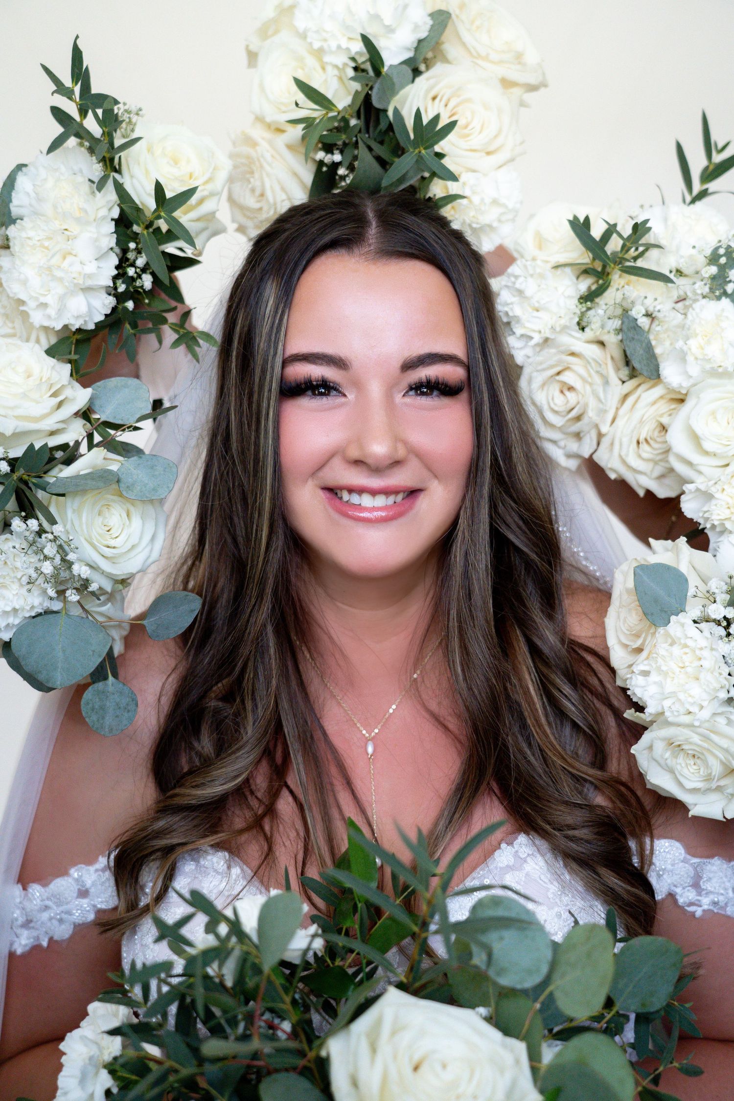 Beautiful bridal portrait with white roses and eucalyptus floral backdrop, wearing off-shoulder lace gown and delicate necklace.