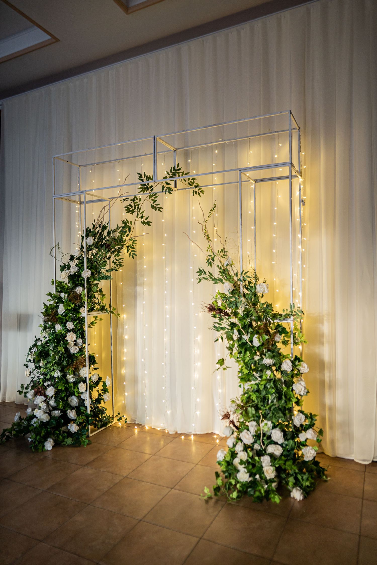 White curtain backdrop with twinkling lights and cascading greenery floral arrangements creating an elegant wedding ceremony setup.