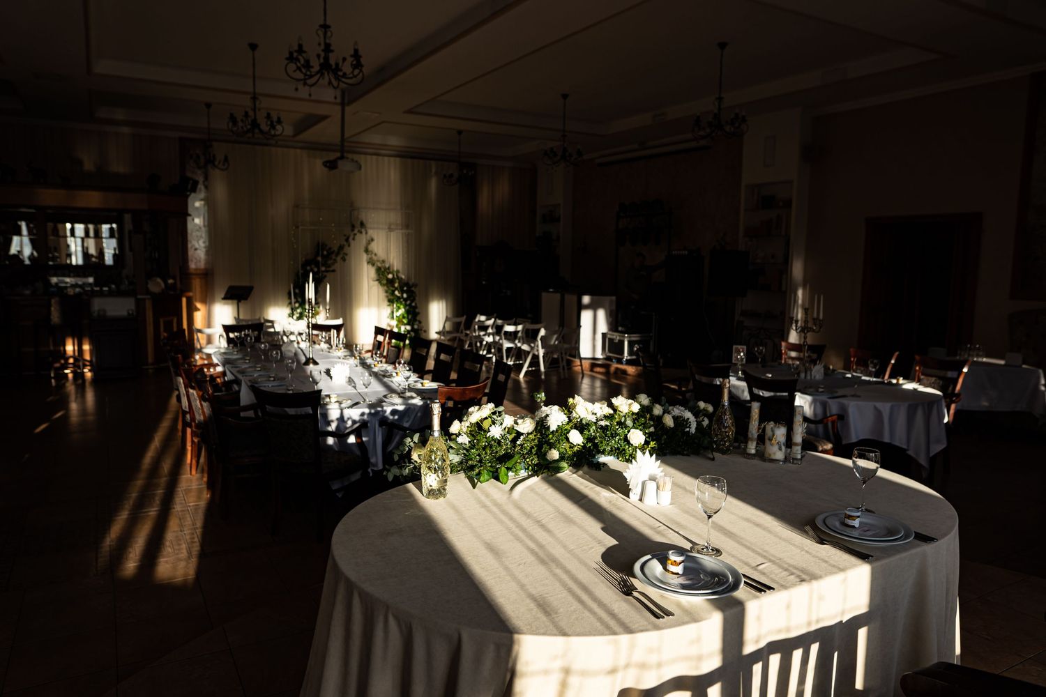 A dimly lit banquet hall with elegant table settings and chandeliers casting a warm glow throughout the space.