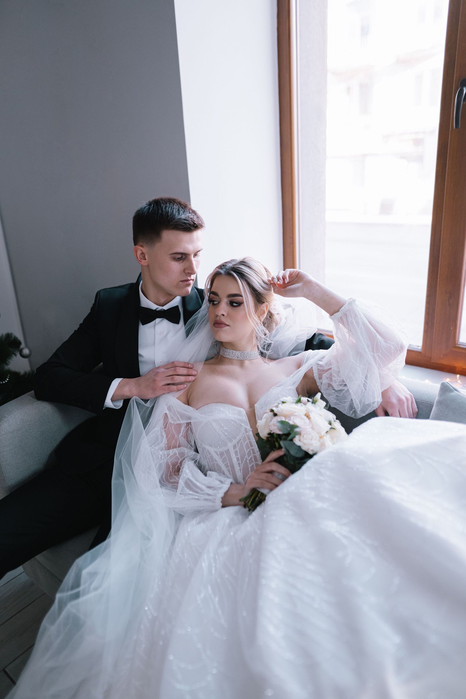 A bride in a white strapless gown and veil lounges with her groom in formal attire near a window.